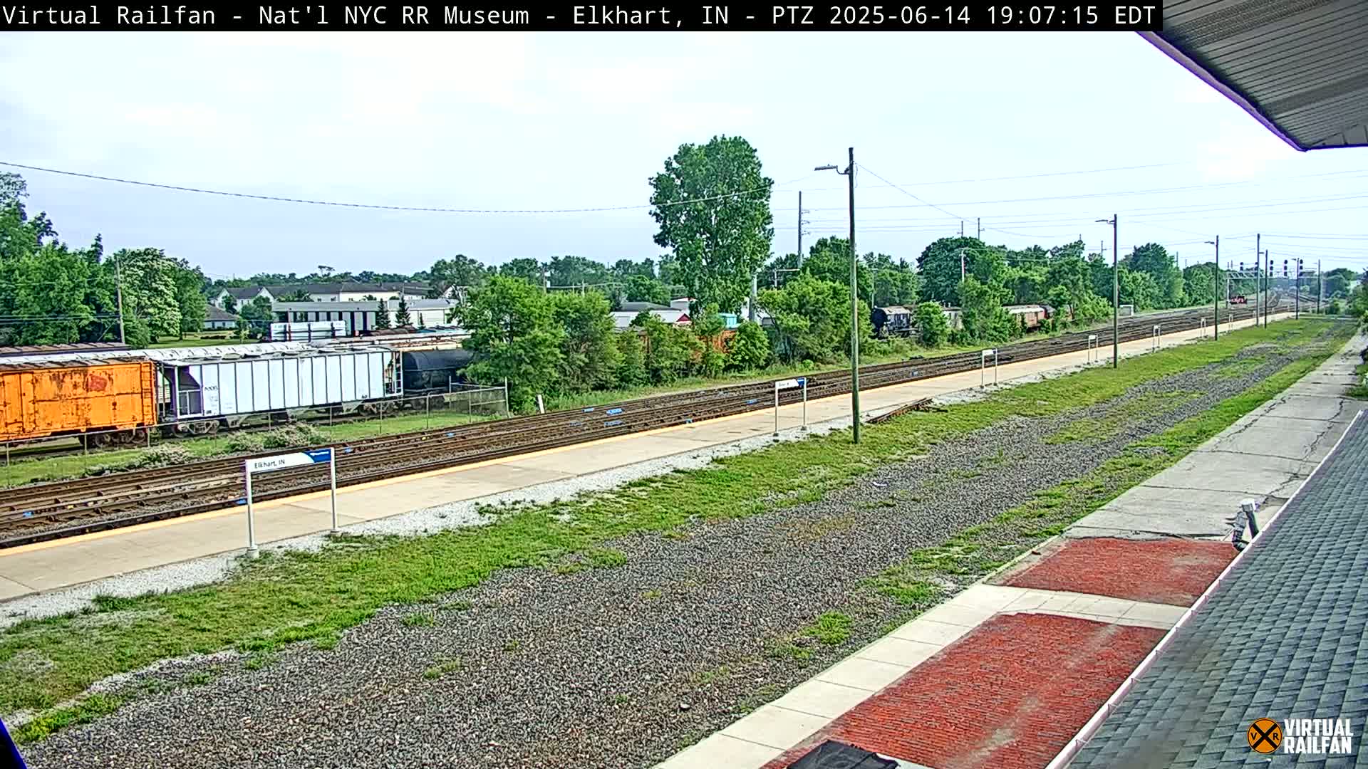 A train station platform with multiple train tracks, some rail cars, and lush green vegetation under a partly cloudy sky.