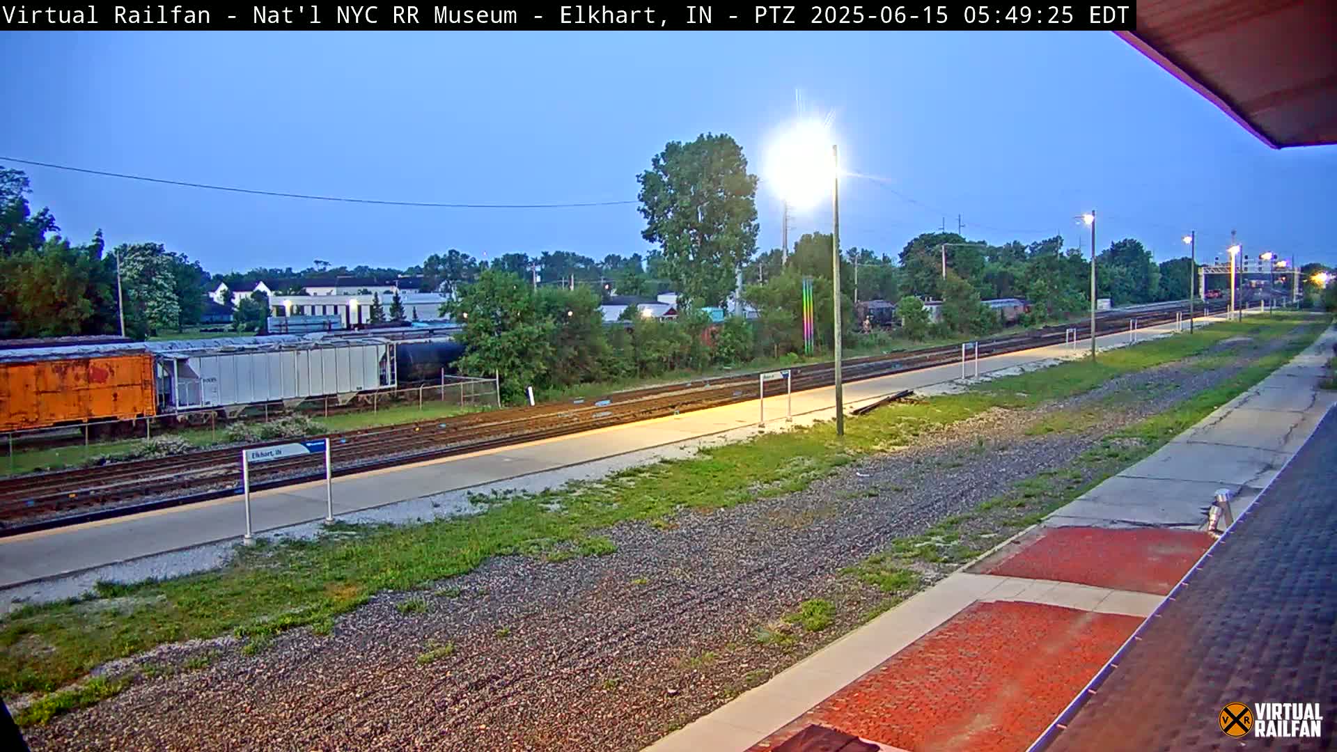 A twilight view of a train station platform with several railway cars and tracks, and grassy areas visible under a clear sky.