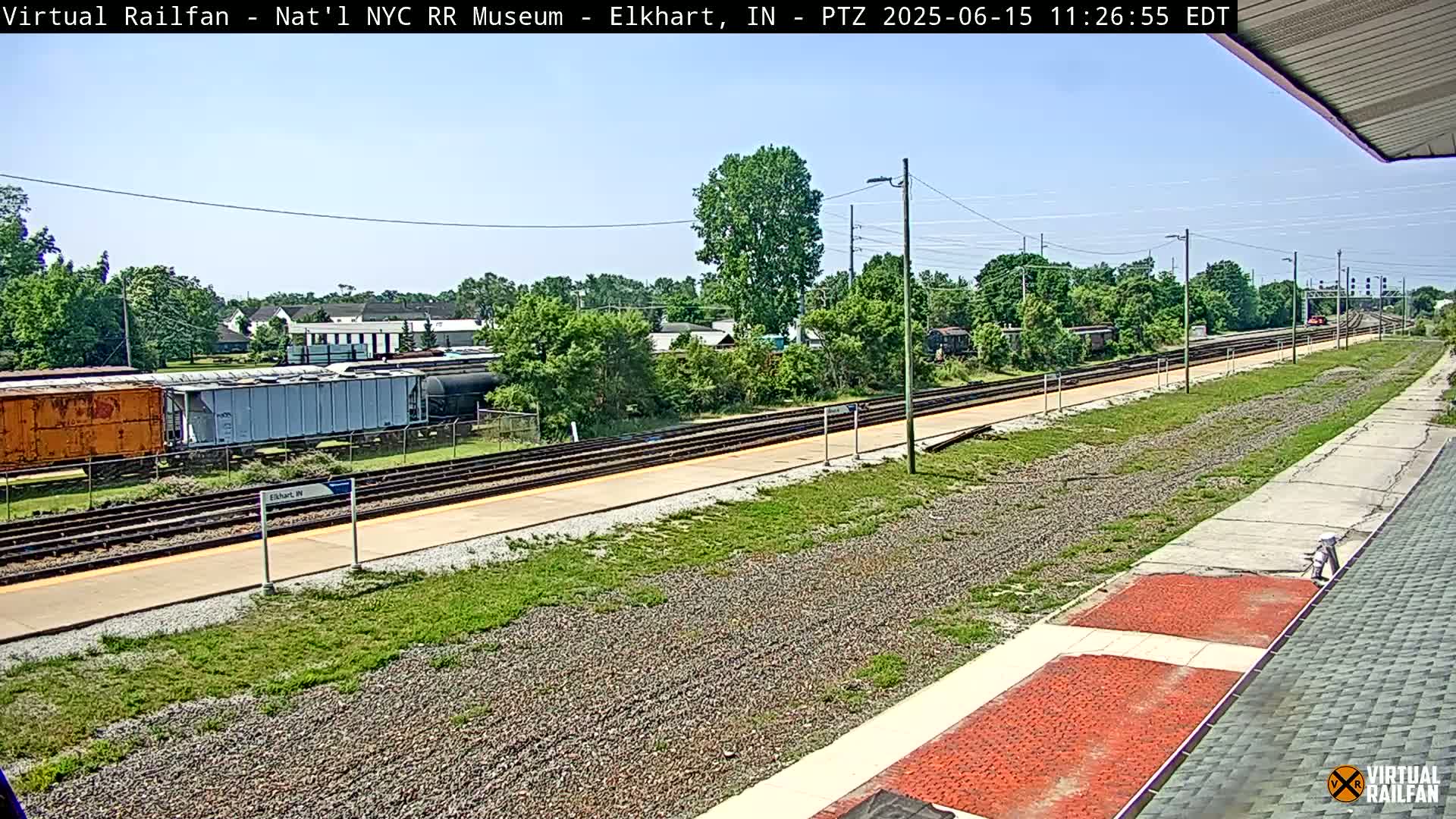 A sunny day reveals a train station platform with several train cars visible in the distance, surrounded by greenery and buildings.