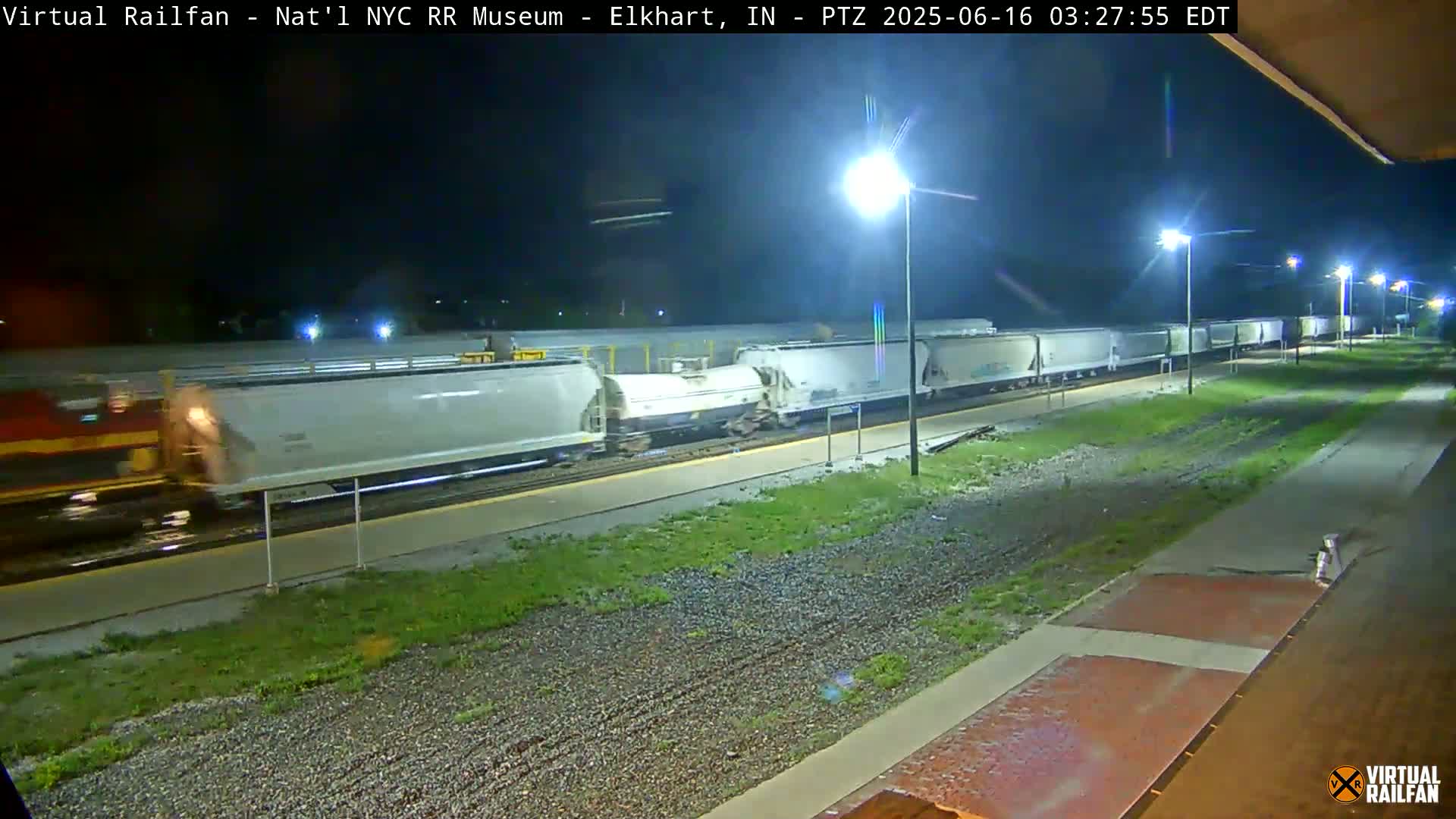 A long freight train of light-grey cars is parked at night next to a platform under several bright lights.