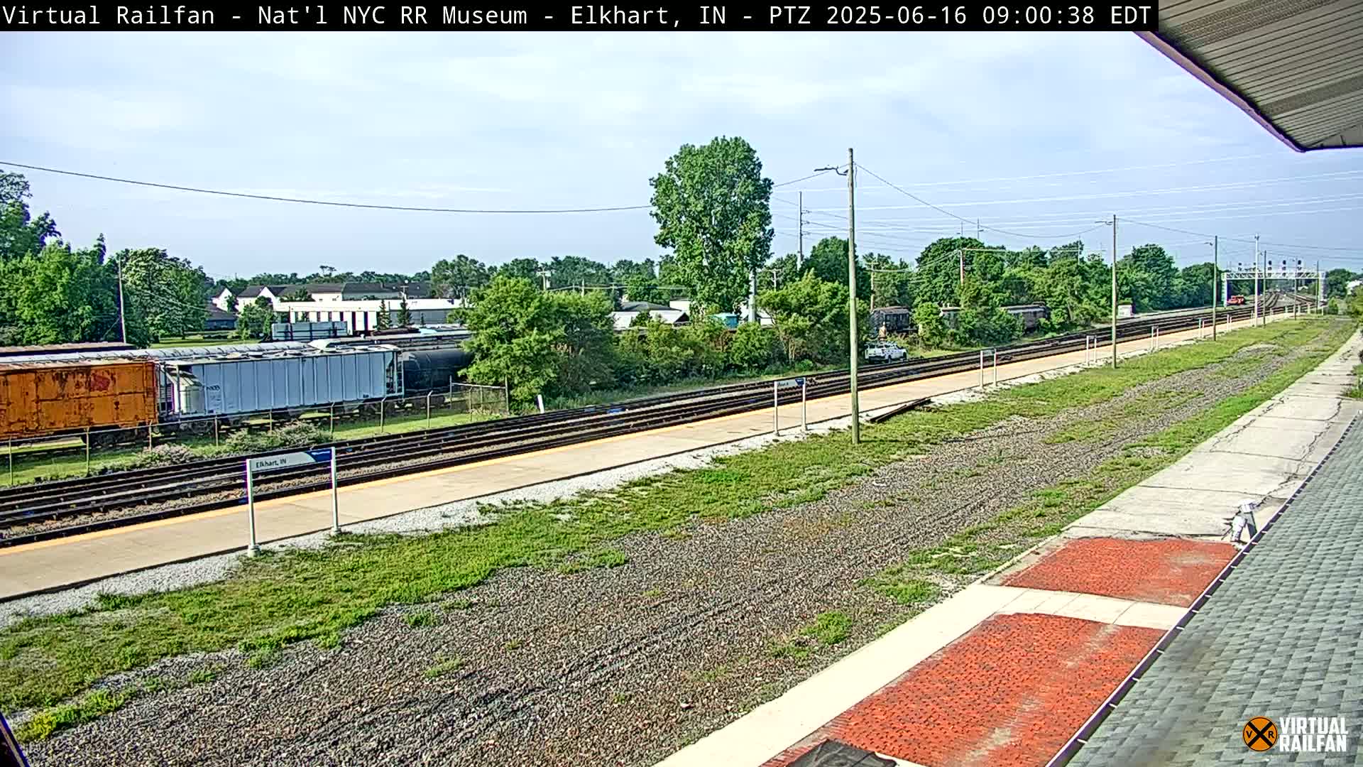 A mostly sunny day at a train station shows railway tracks with several freight cars and a platform, surrounded by green vegetation and some buildings in the distance.