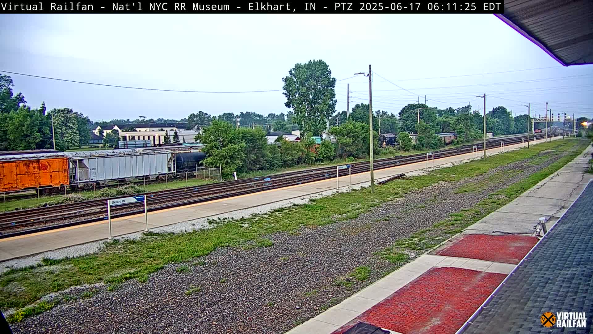 A train station platform with several railway tracks, freight cars, and overgrown vegetation, under a partly cloudy sky.
