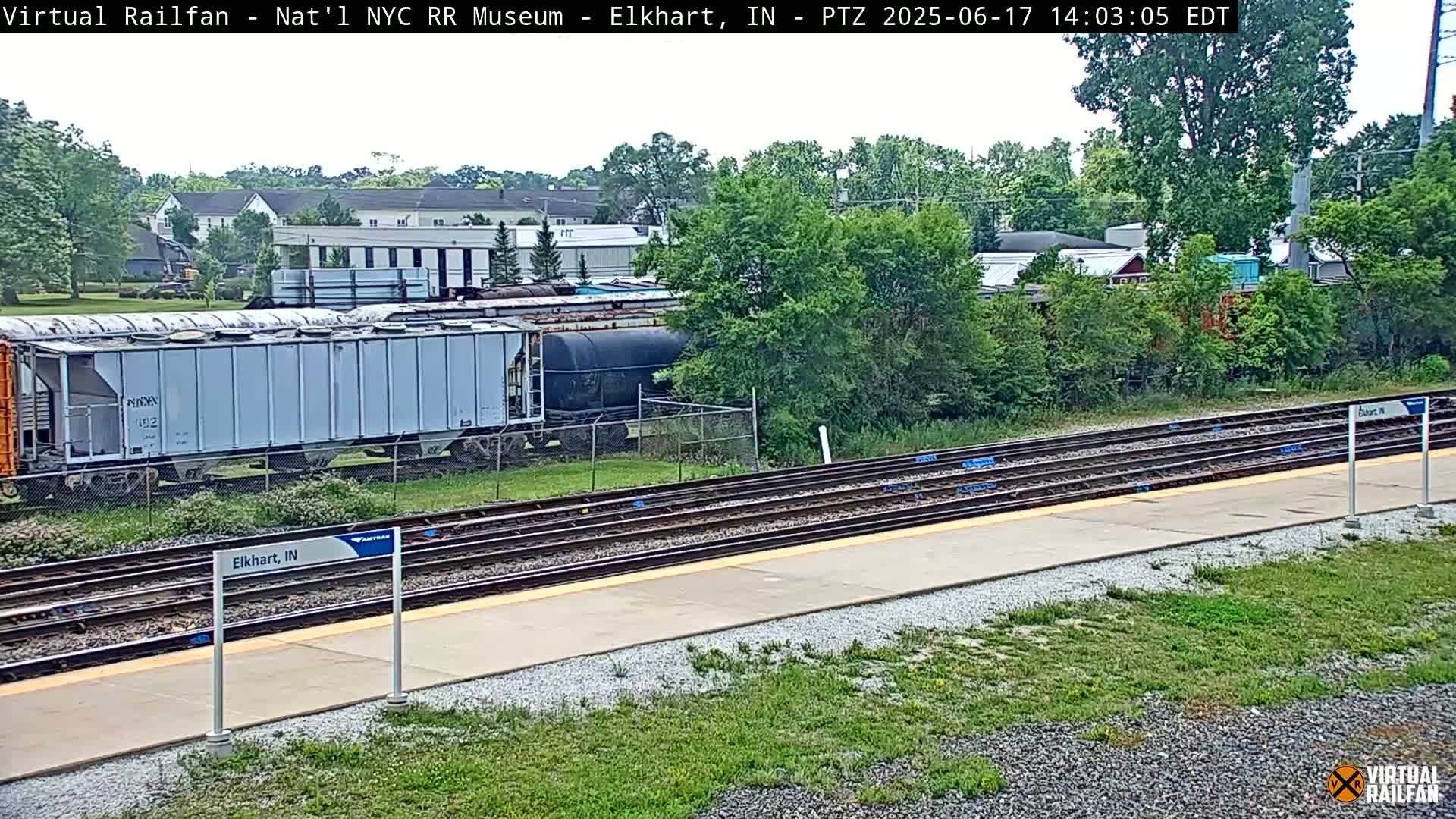 A train of boxcars and tankers sits on a railway beside a platform on a partly sunny day.