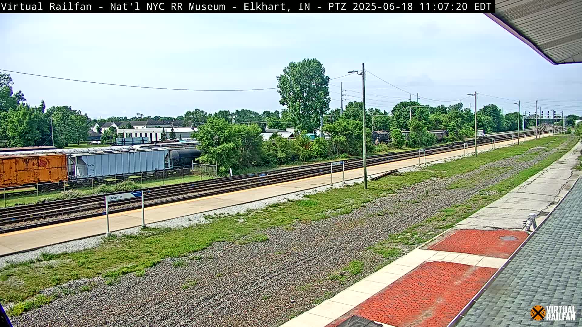Under a mostly clear sky, a train station platform with several rail lines stretches into the distance, past some parked train cars and greenery.
