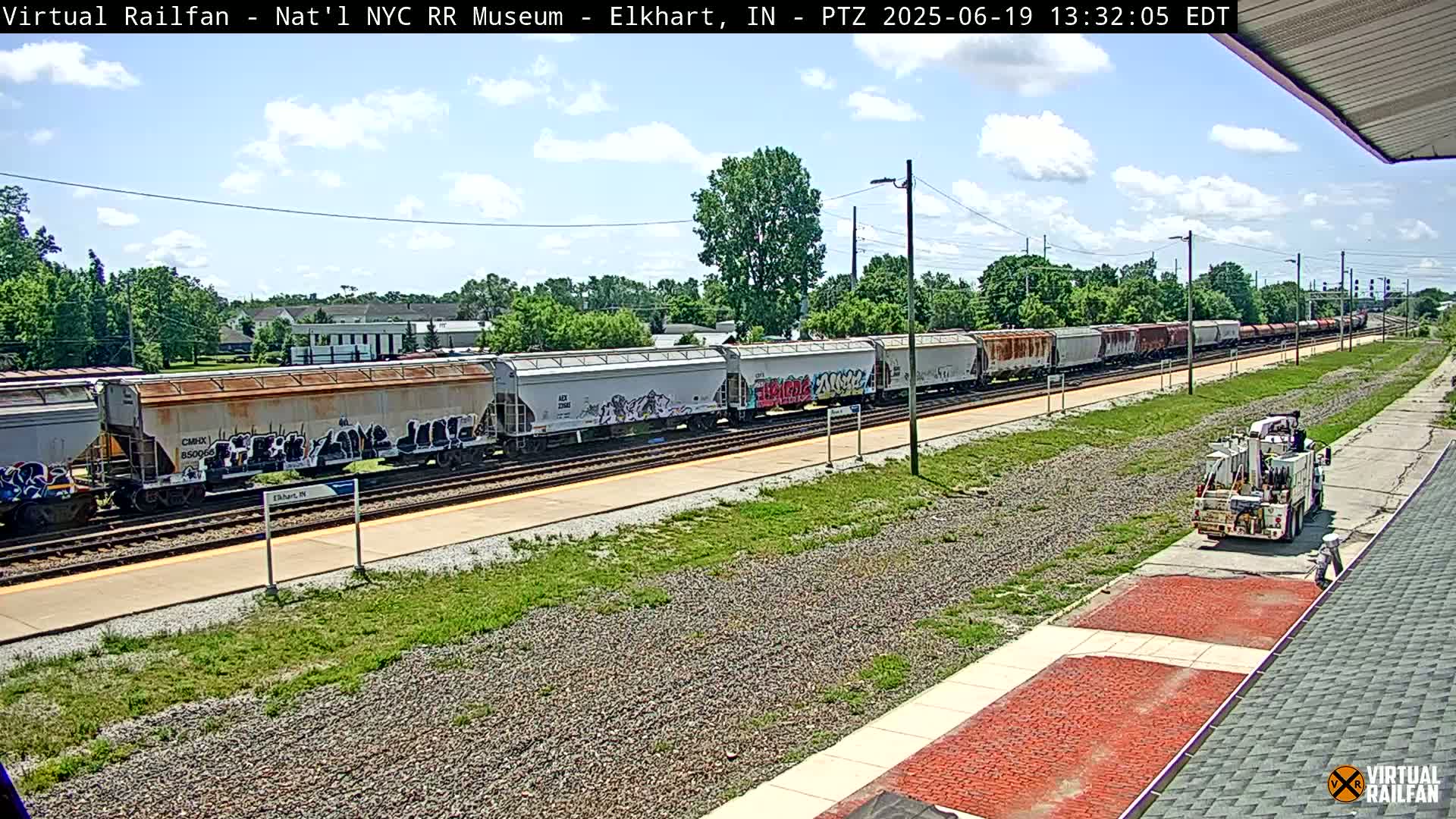 A long freight train with graffiti on some of the cars is visible on a sunny day, next to a maintenance truck and a paved walkway.