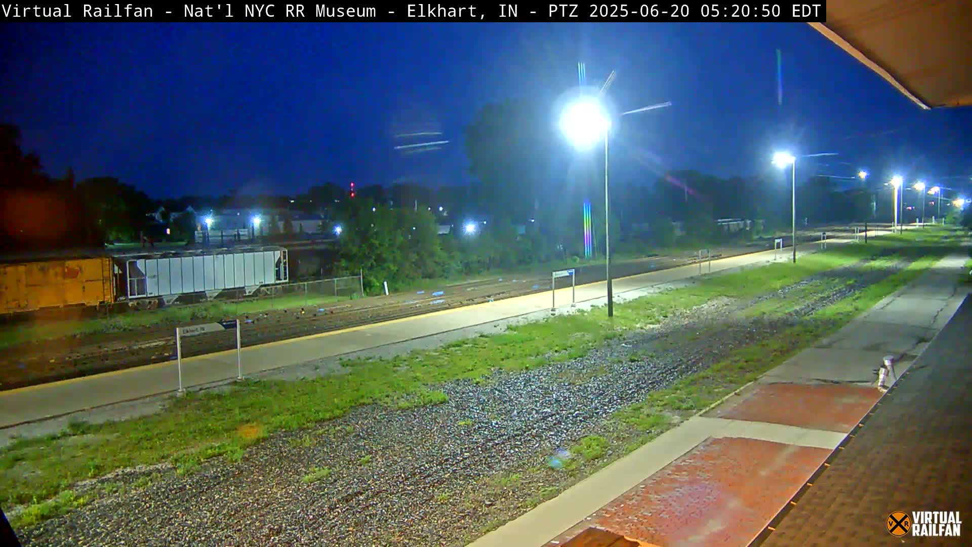 A nighttime view of a train station platform with railway tracks and parked railcars, illuminated by streetlights under a dark blue sky.
