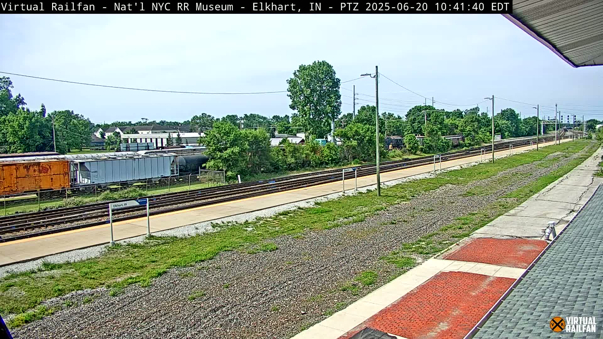 A sunny day at a train station shows multiple train cars parked on parallel tracks, with a platform and grassy areas surrounding the tracks.