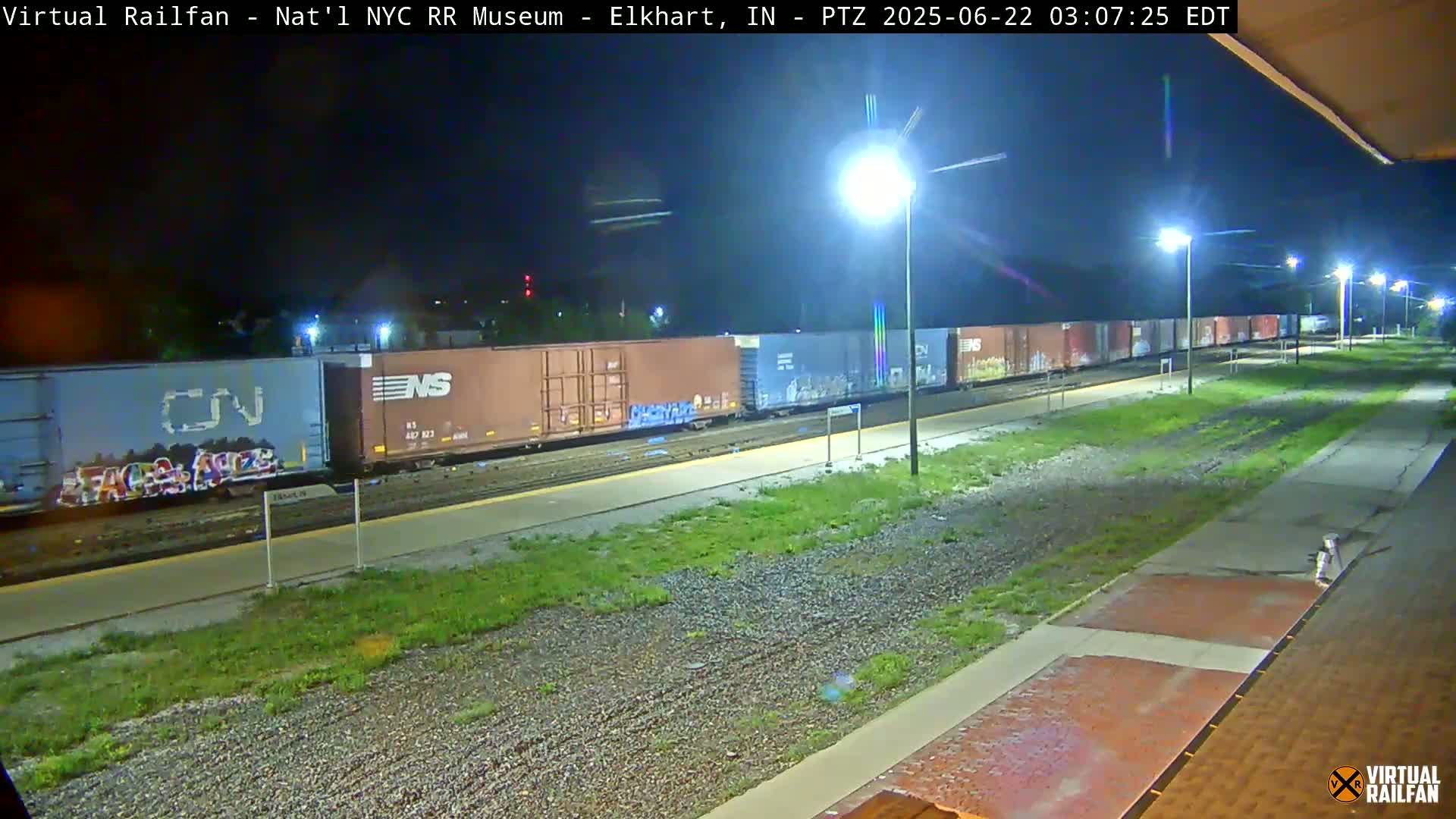 A long freight train sits at night on railway tracks beside a grassy area illuminated by several streetlights.