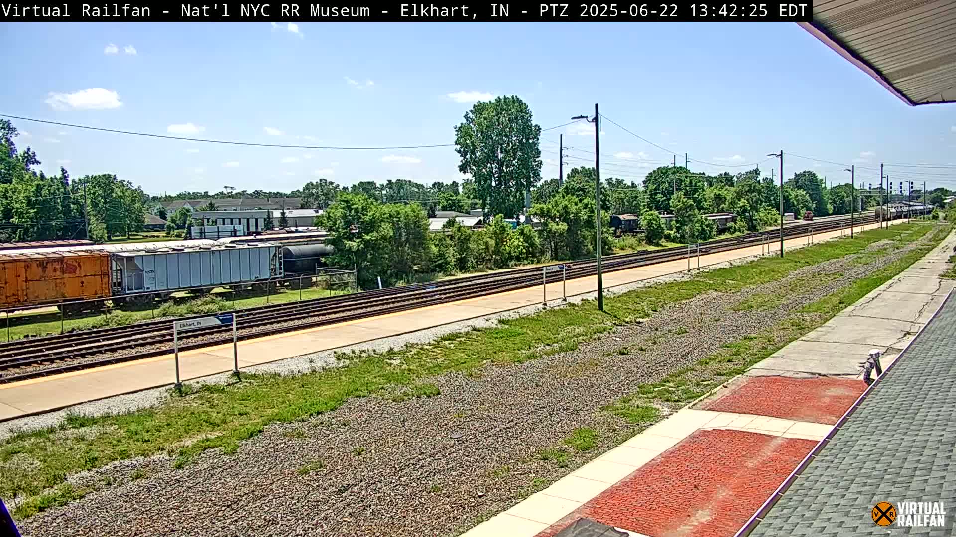 A sunny day at a train station shows multiple sets of train tracks extending into the distance, lined with grassy areas and several railway cars.