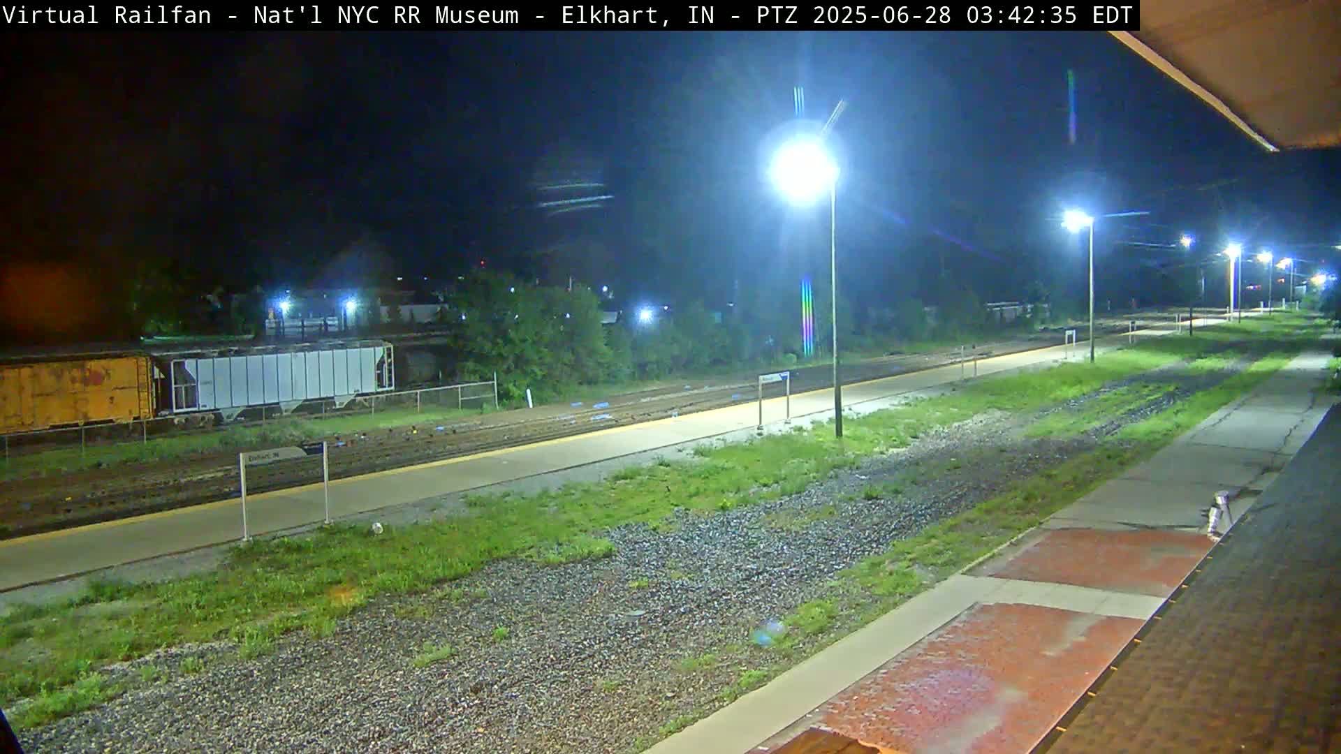 A nighttime view of a train station platform and railway tracks, with several railcars visible in the distance under nighttime lighting conditions.