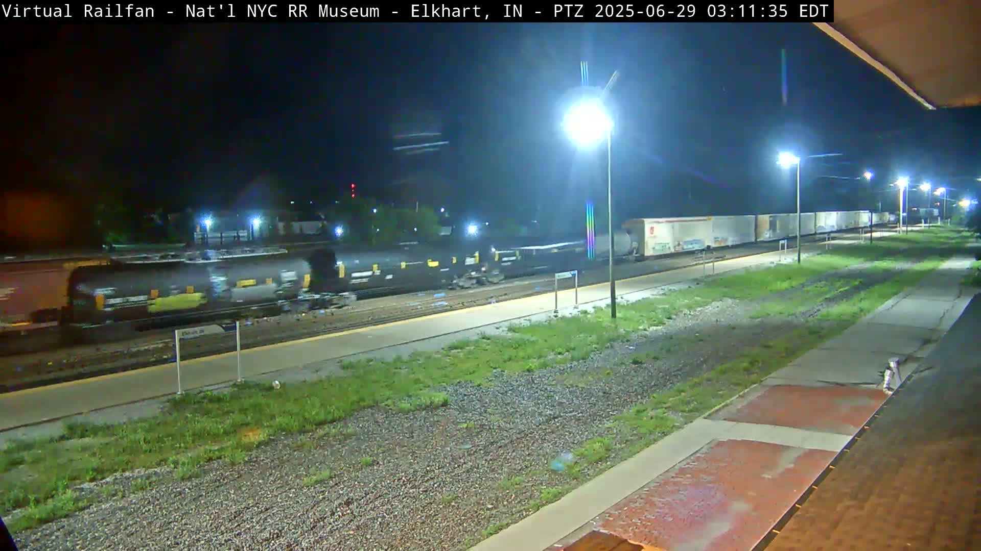 A long freight train passes under nighttime lighting at a station with grassy areas beside the tracks.
