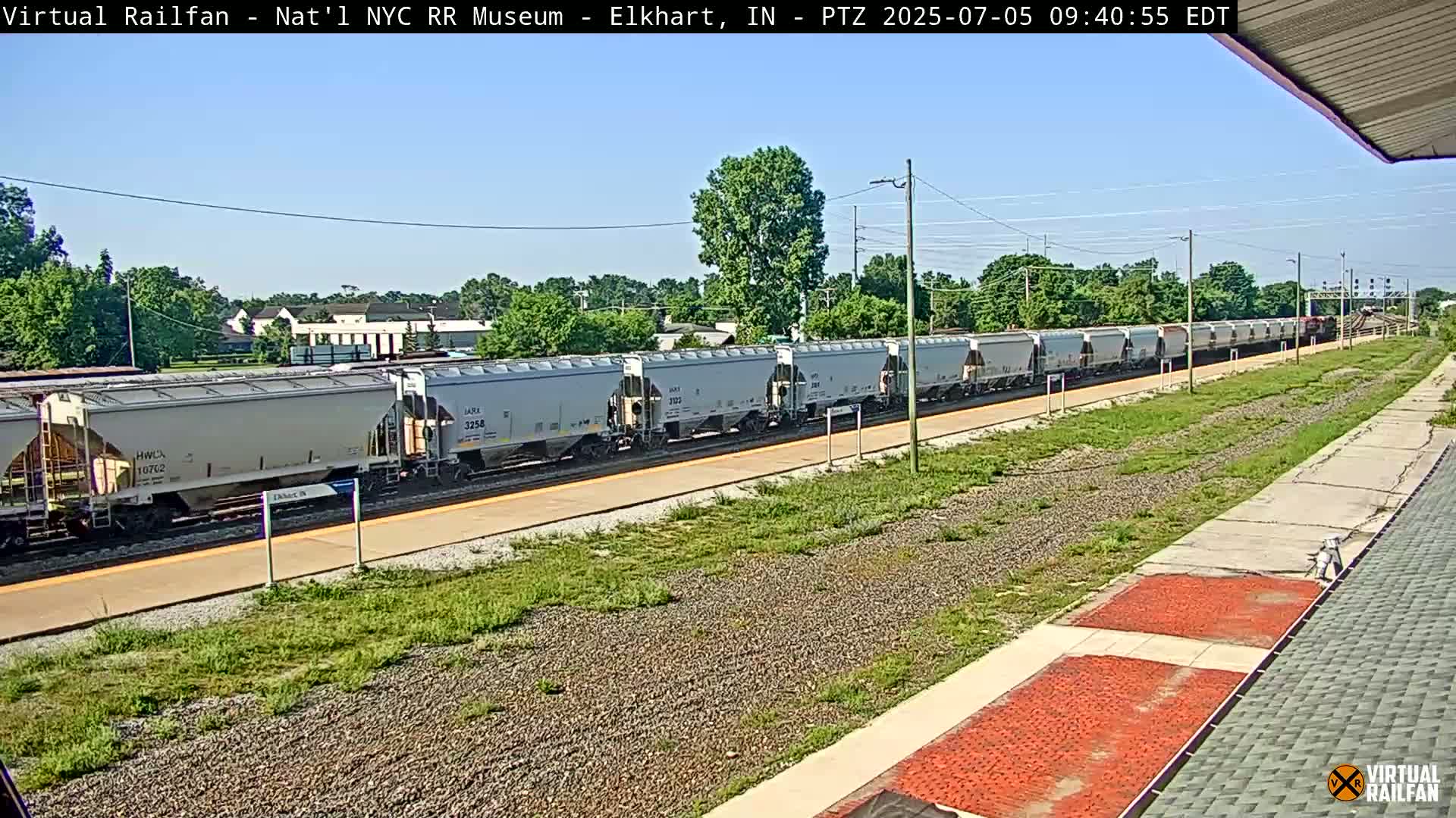 A long freight train of light gray covered hopper cars is seen passing by a train platform on a sunny day.