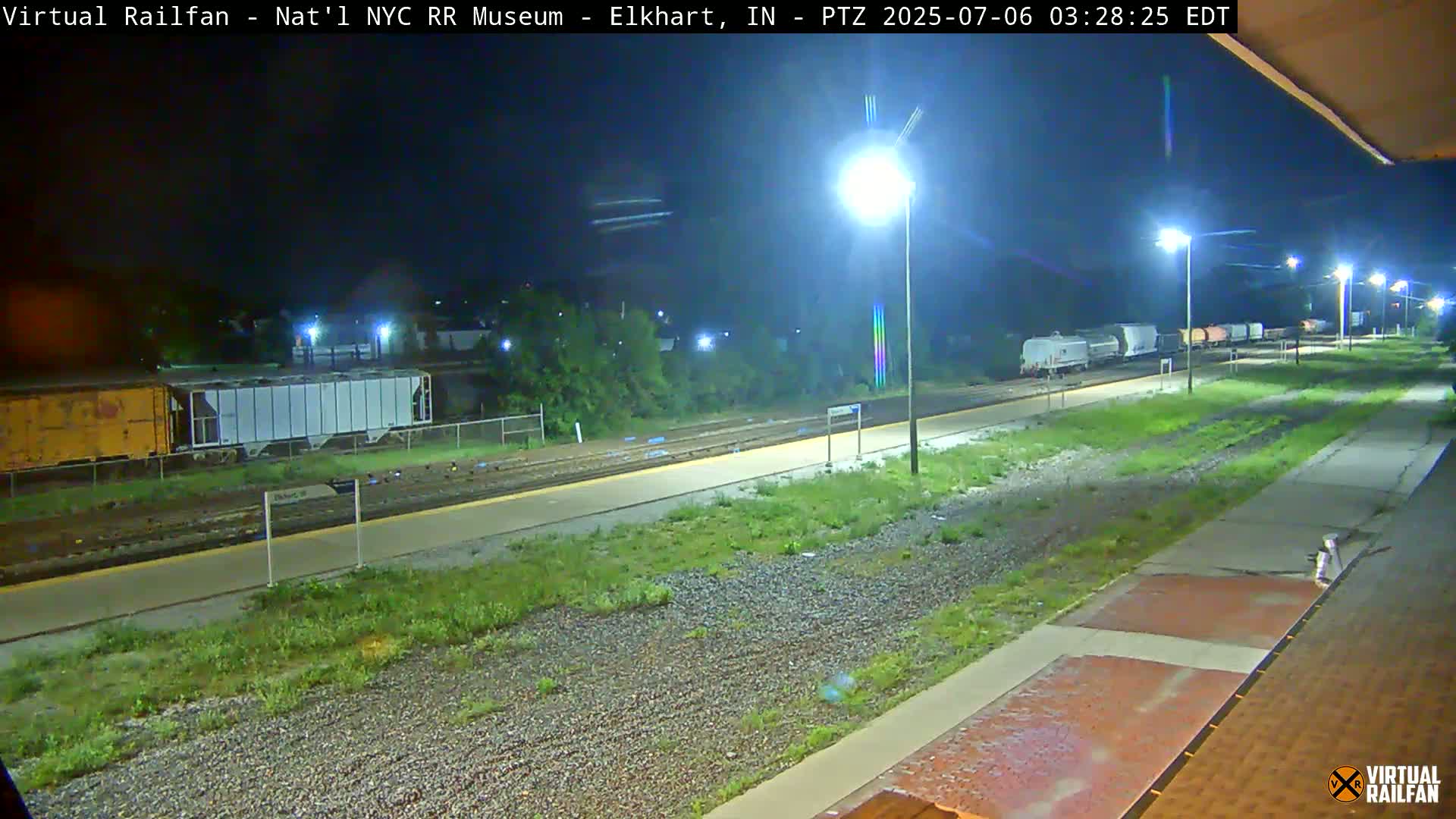 A nighttime view of a train station platform with a freight train parked on the tracks, illuminated by several streetlights.