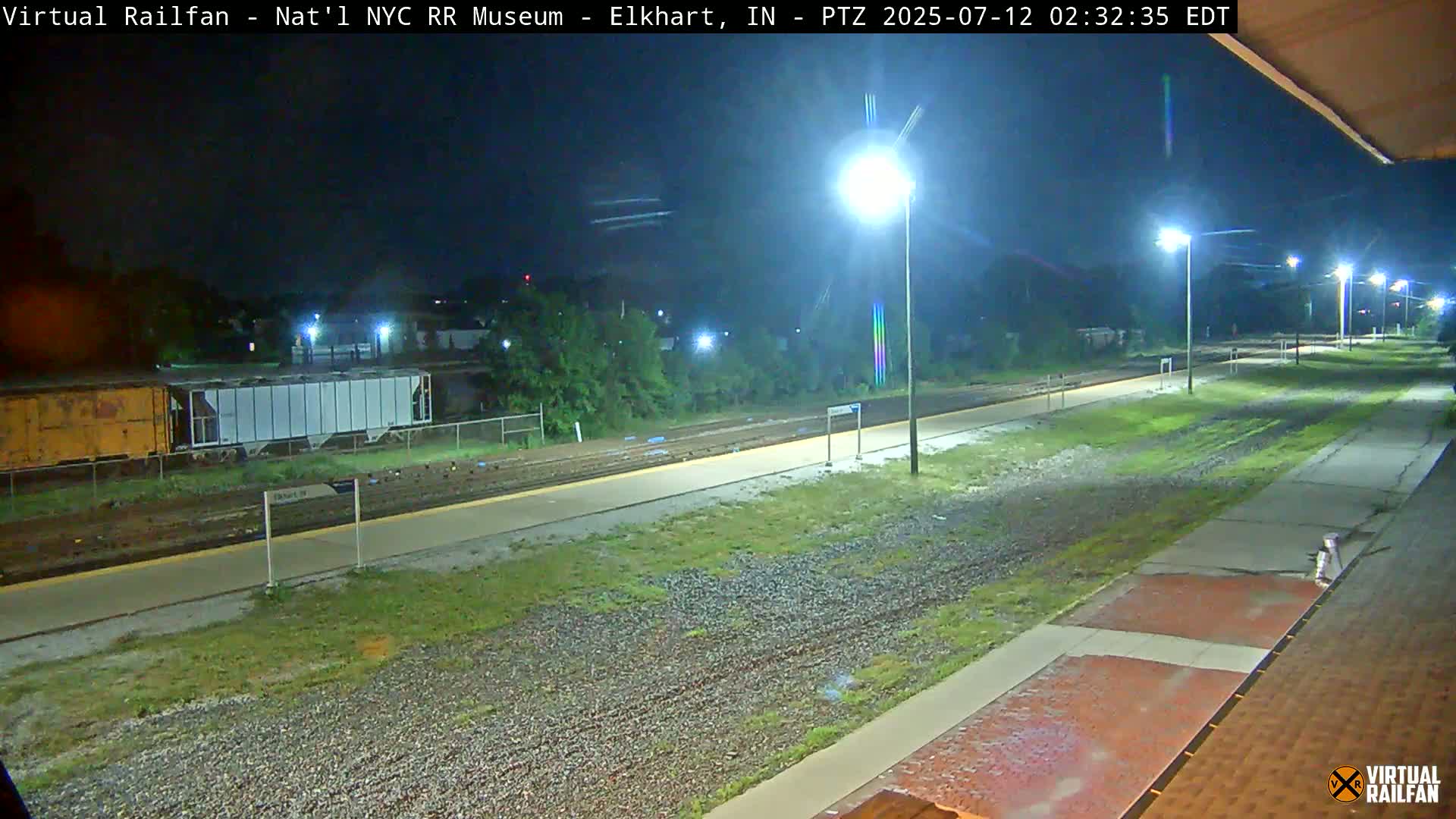 A nighttime view of a train station platform with a freight train parked on the tracks, illuminated by several streetlights.