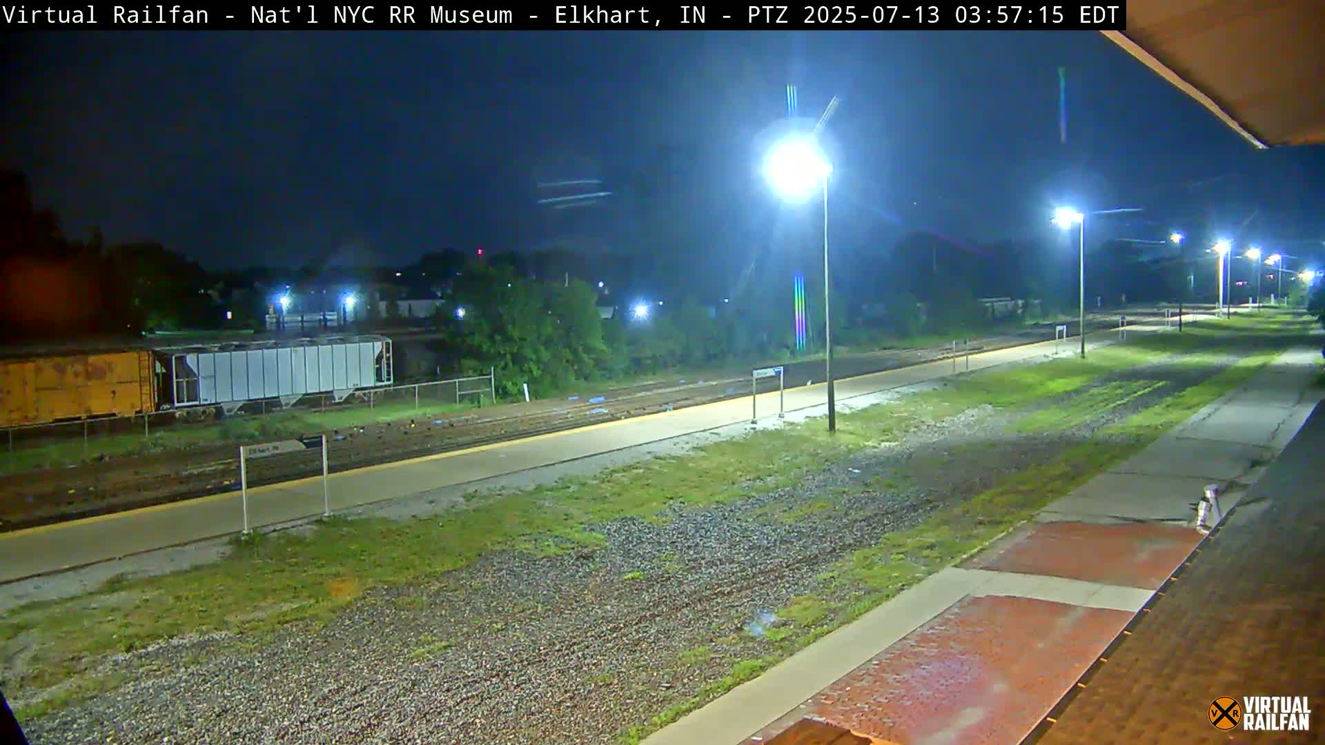 A nighttime view of a train station platform and railway tracks, illuminated by several streetlights.