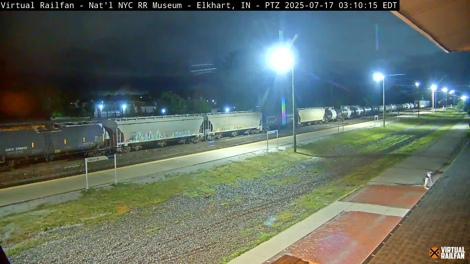 A long freight train sits parked at night under bright lights next to a grassy area.