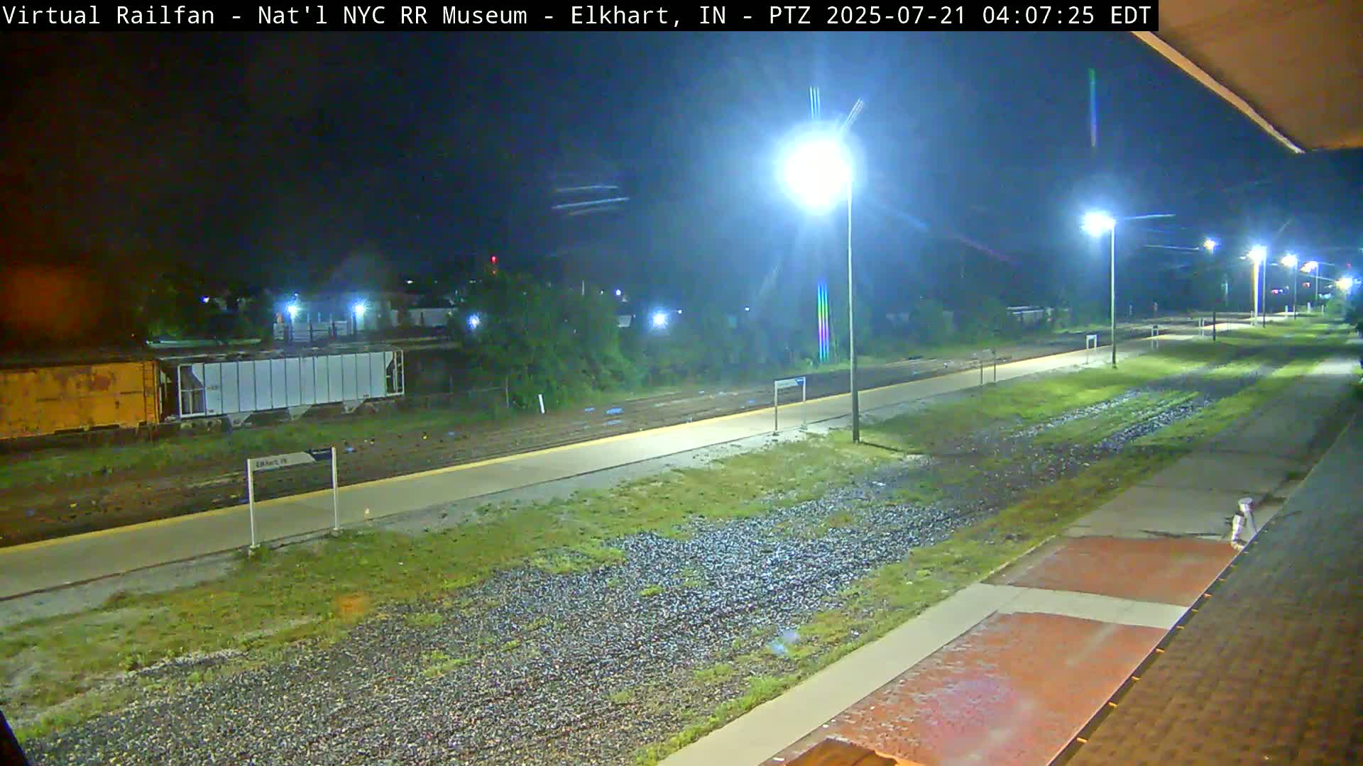 A nighttime view of a train platform and tracks, illuminated by bright lights, with a few train cars visible in the distance.