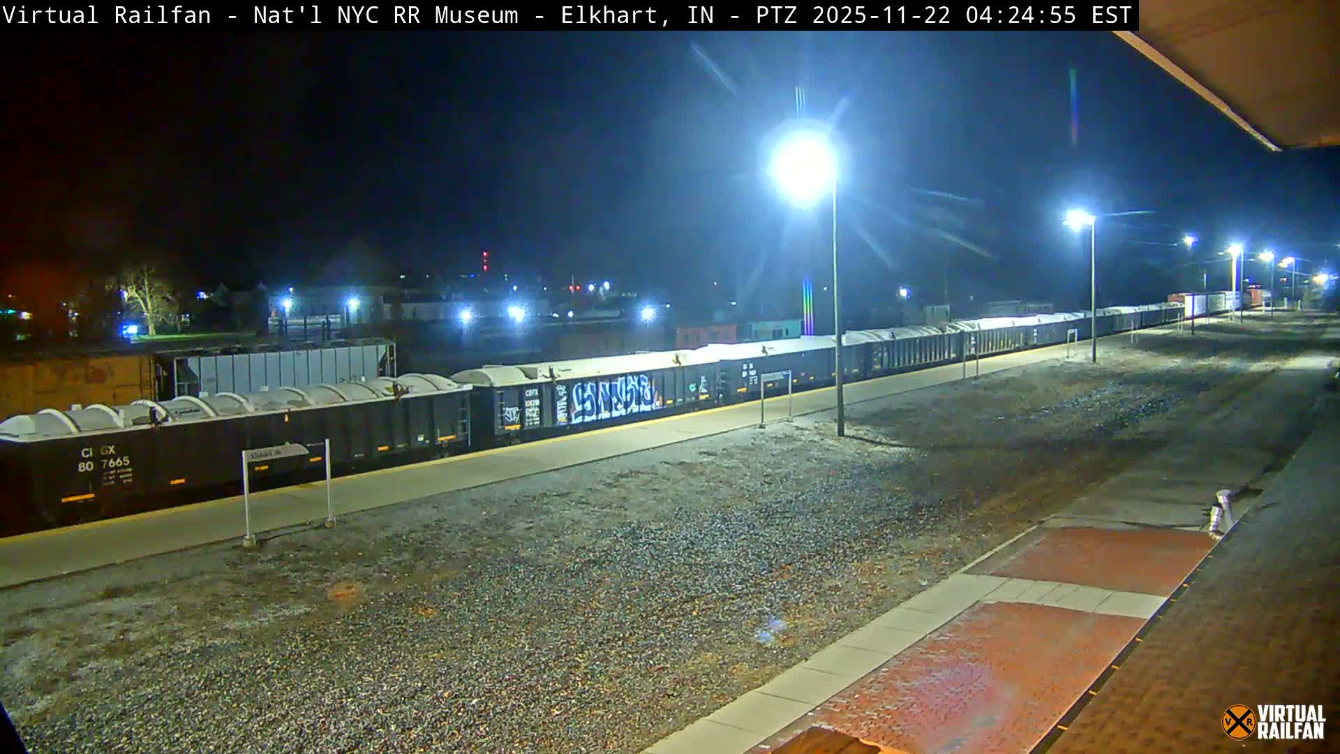 A long freight train with covered hopper cars, some bearing graffiti, is parked on tracks next to a brightly lit station platform at night, where the ground and platform surfaces appear damp.