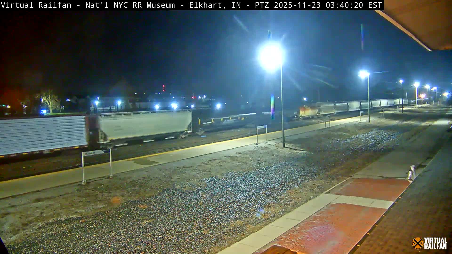 A long freight train with numerous white container cars moves on multiple tracks next to a station platform at night, illuminated by bright streetlights under clear but damp conditions.