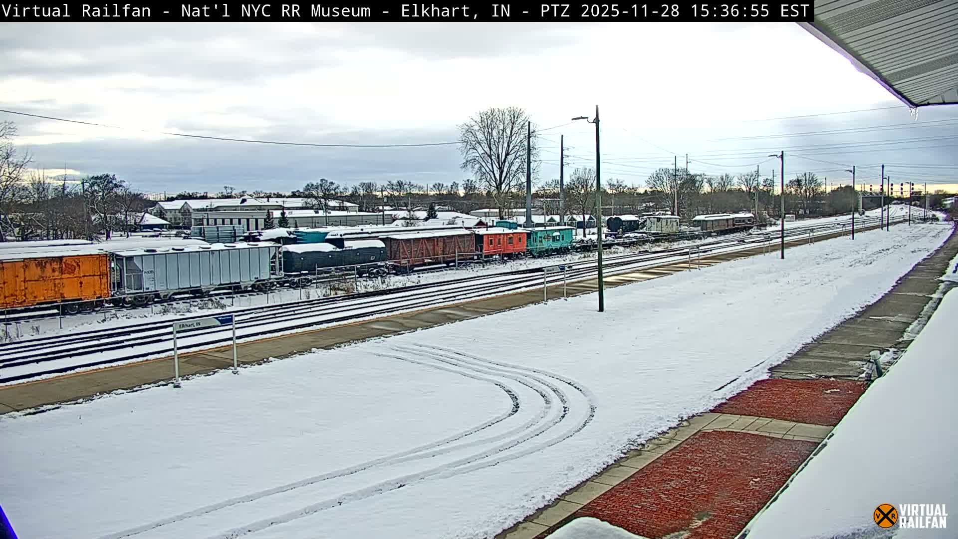A snowy winter day depicts a wide view of multiple train tracks and an array of colorful train cars parked in a railway yard, with a snow-covered platform in the foreground and overcast skies above.