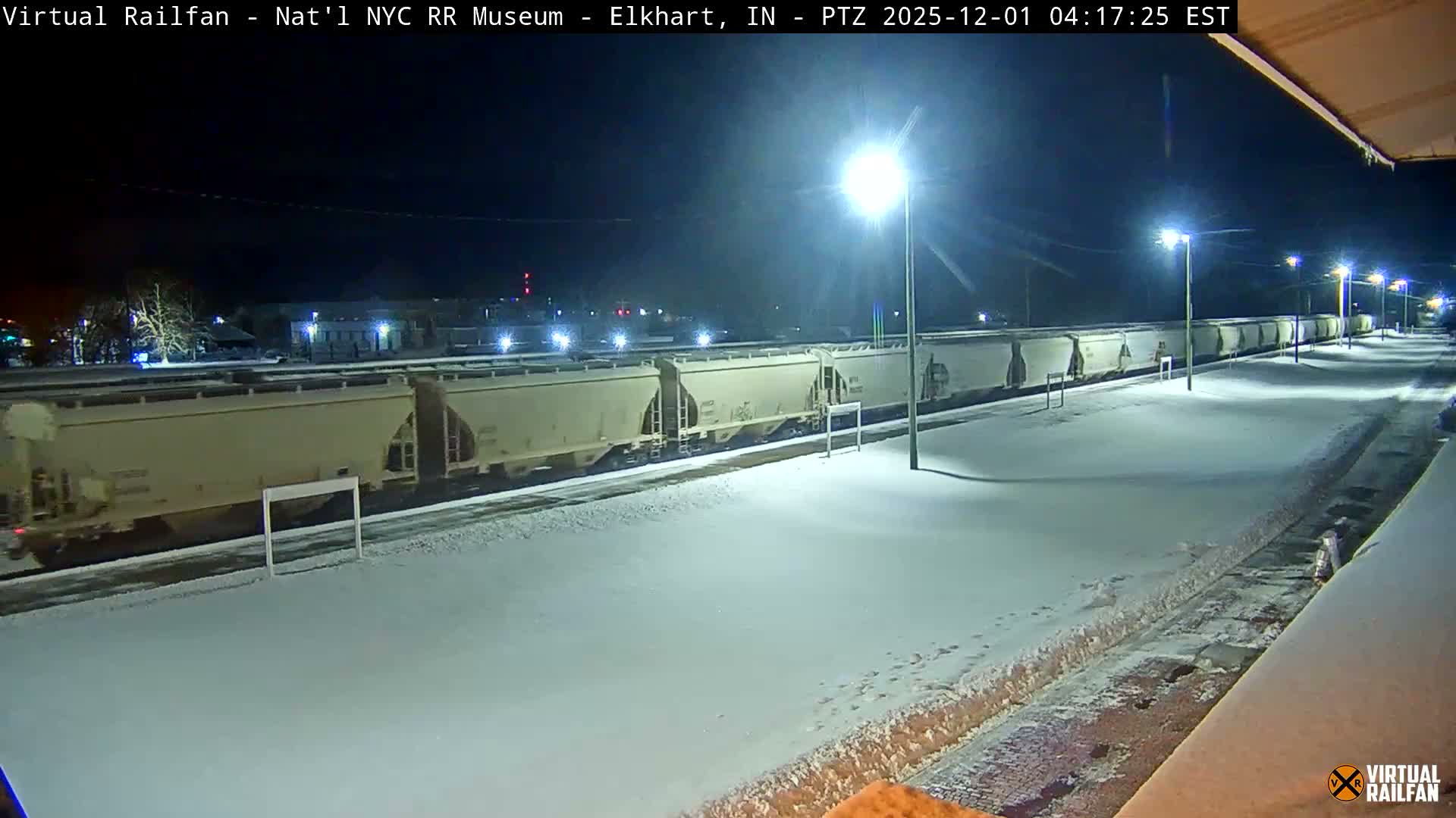 A long train of beige hopper cars sits on snow-covered tracks at a rail yard during a clear, cold, and snowy night, illuminated by multiple bright streetlights.