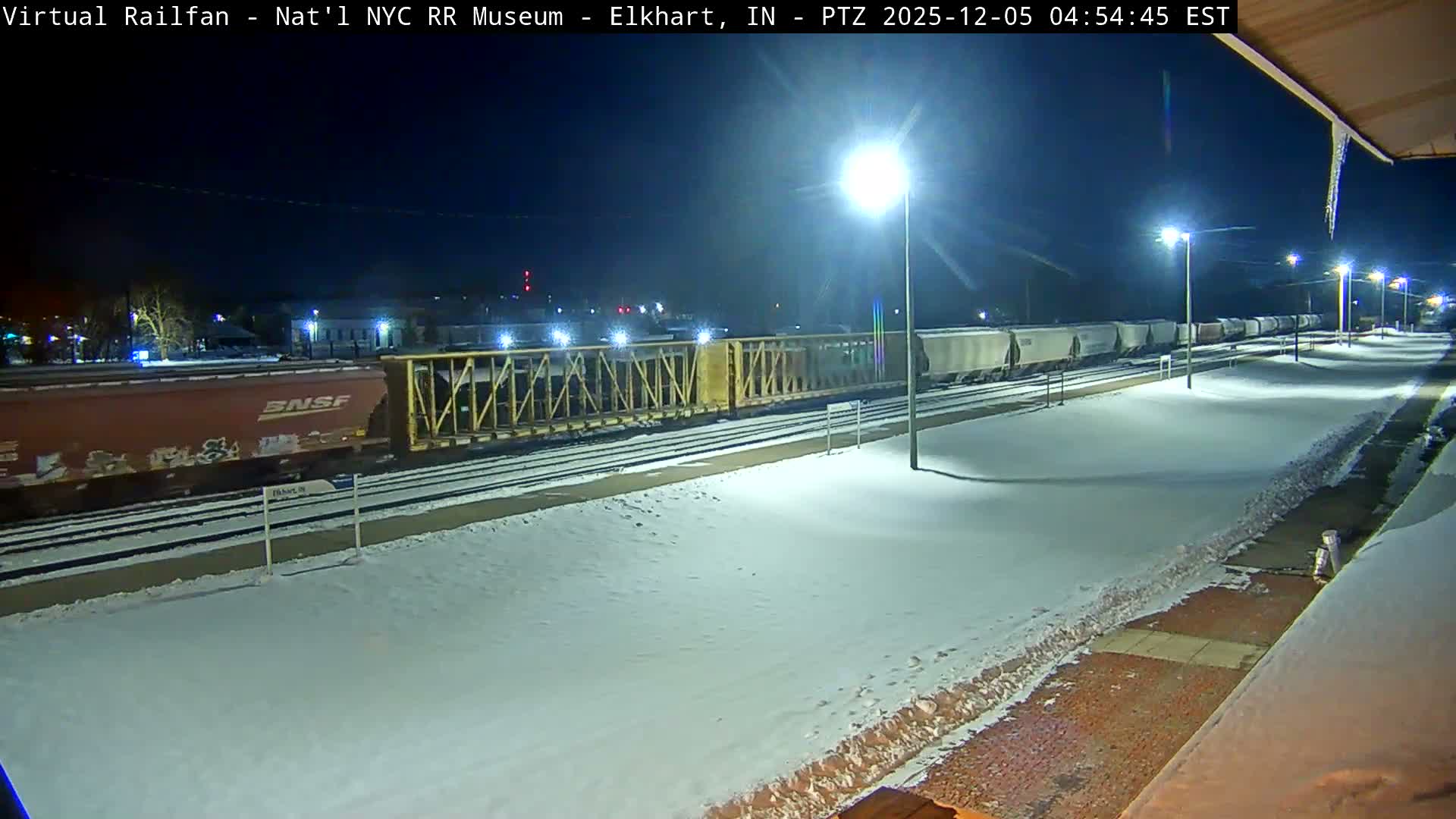 A long freight train sits on tracks beside snow-covered platforms at night, illuminated by multiple streetlights under a clear, dark sky, with icicles visible on an overhang indicating freezing winter conditions.