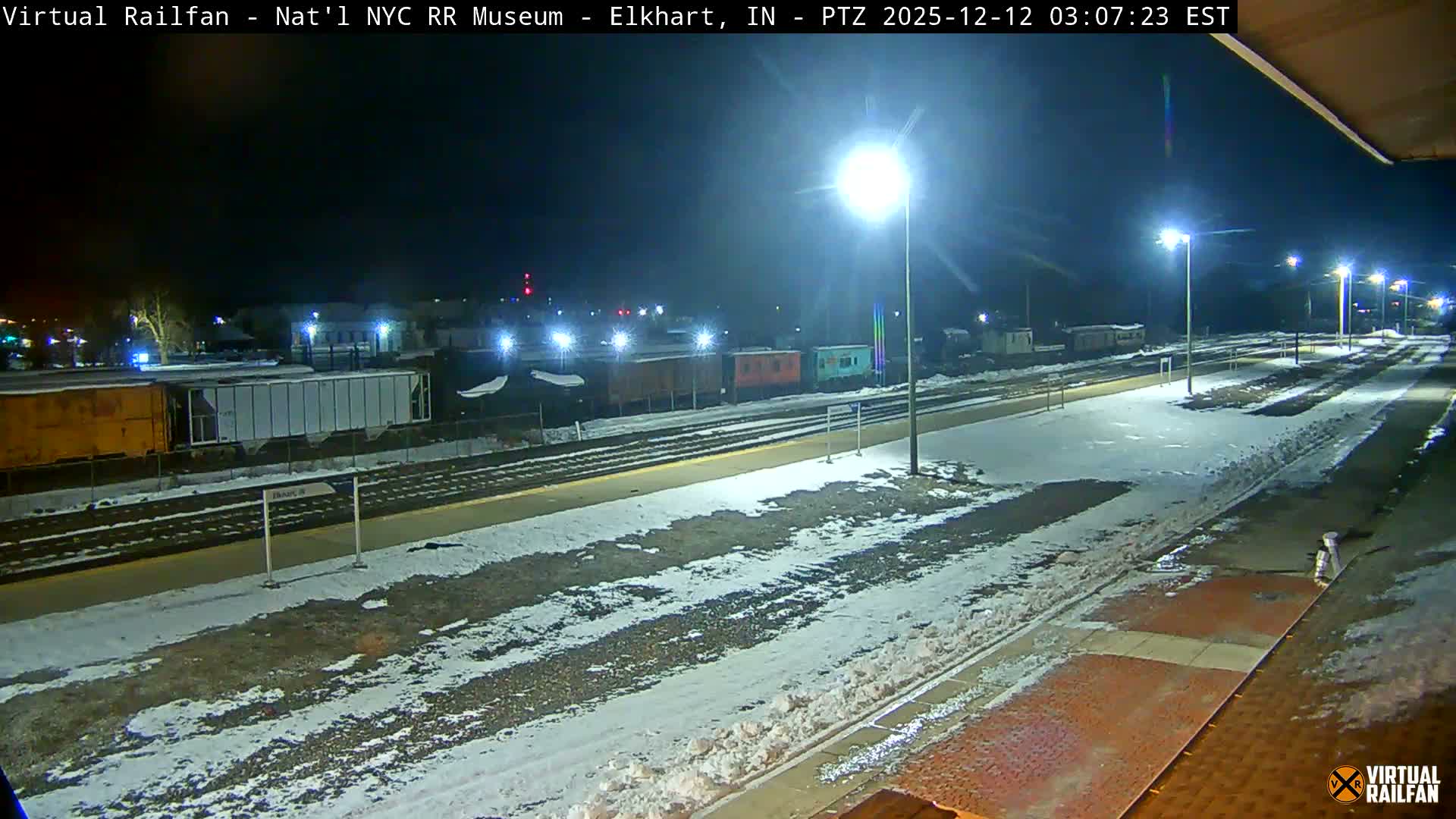 A wide nighttime view of multiple railway tracks and platforms covered in a light layer of snow, with various freight cars visible on the left and bright streetlights illuminating the scene under a dark sky.