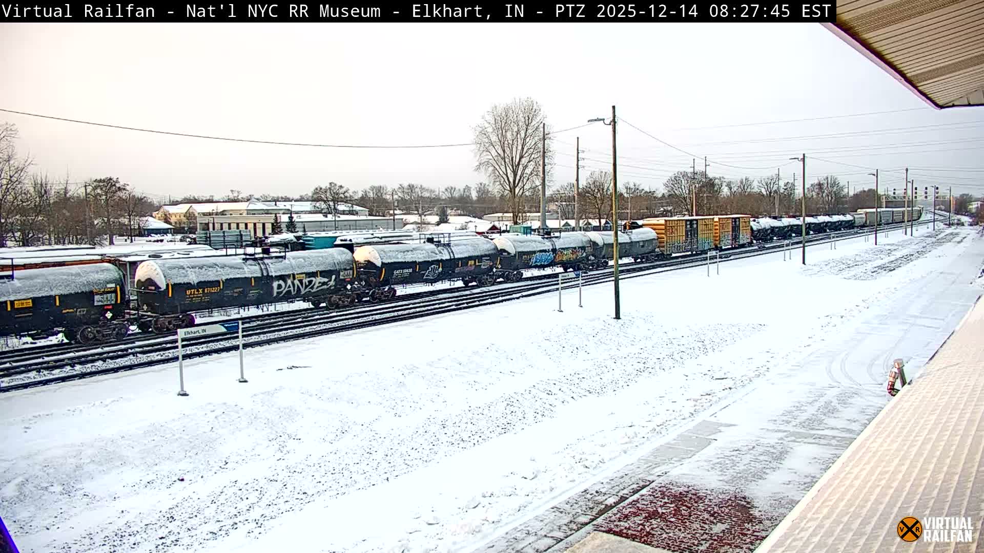 A wide nighttime view of multiple railway tracks and platforms covered in a light layer of snow, with various freight cars visible on the left and bright streetlights illuminating the scene under a dark sky.
