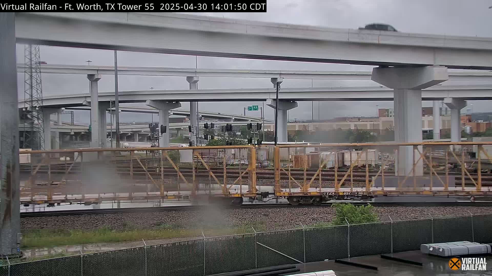 A freight train passes under a large highway overpass in overcast weather.