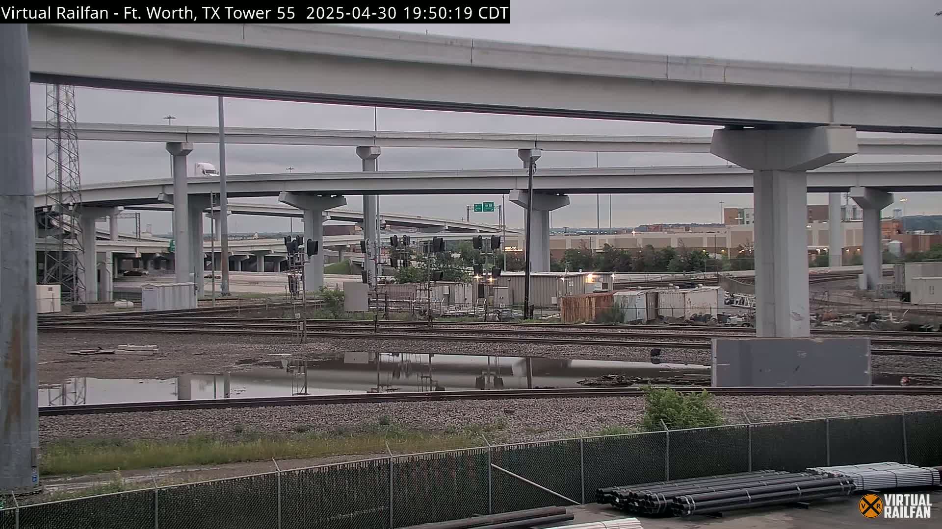A view of an overcast day showing railroad tracks and a large highway overpass with puddles of water in the foreground.