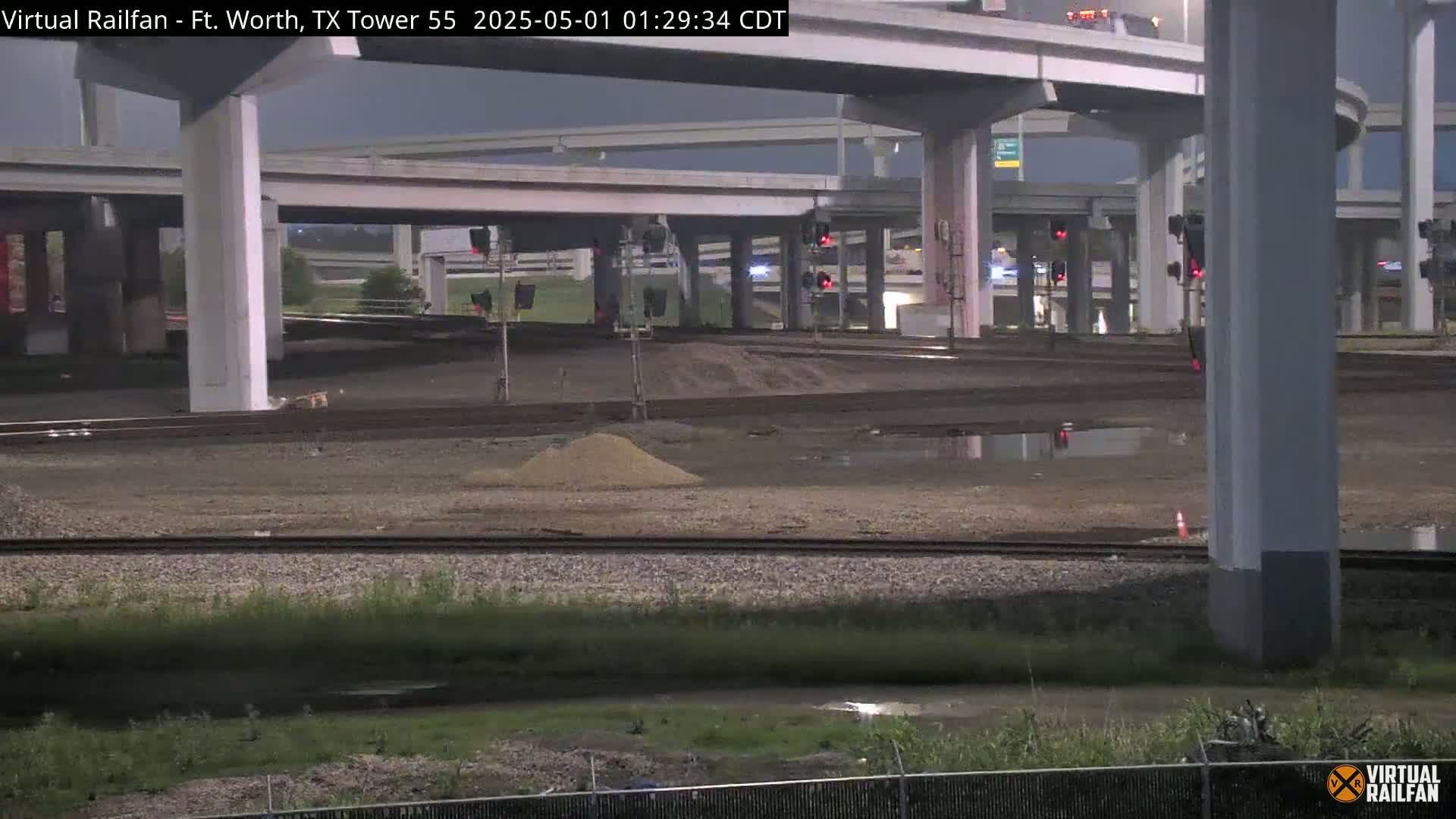 A nighttime view of a railway and highway interchange, with puddles of water on the ground and piles of sand.