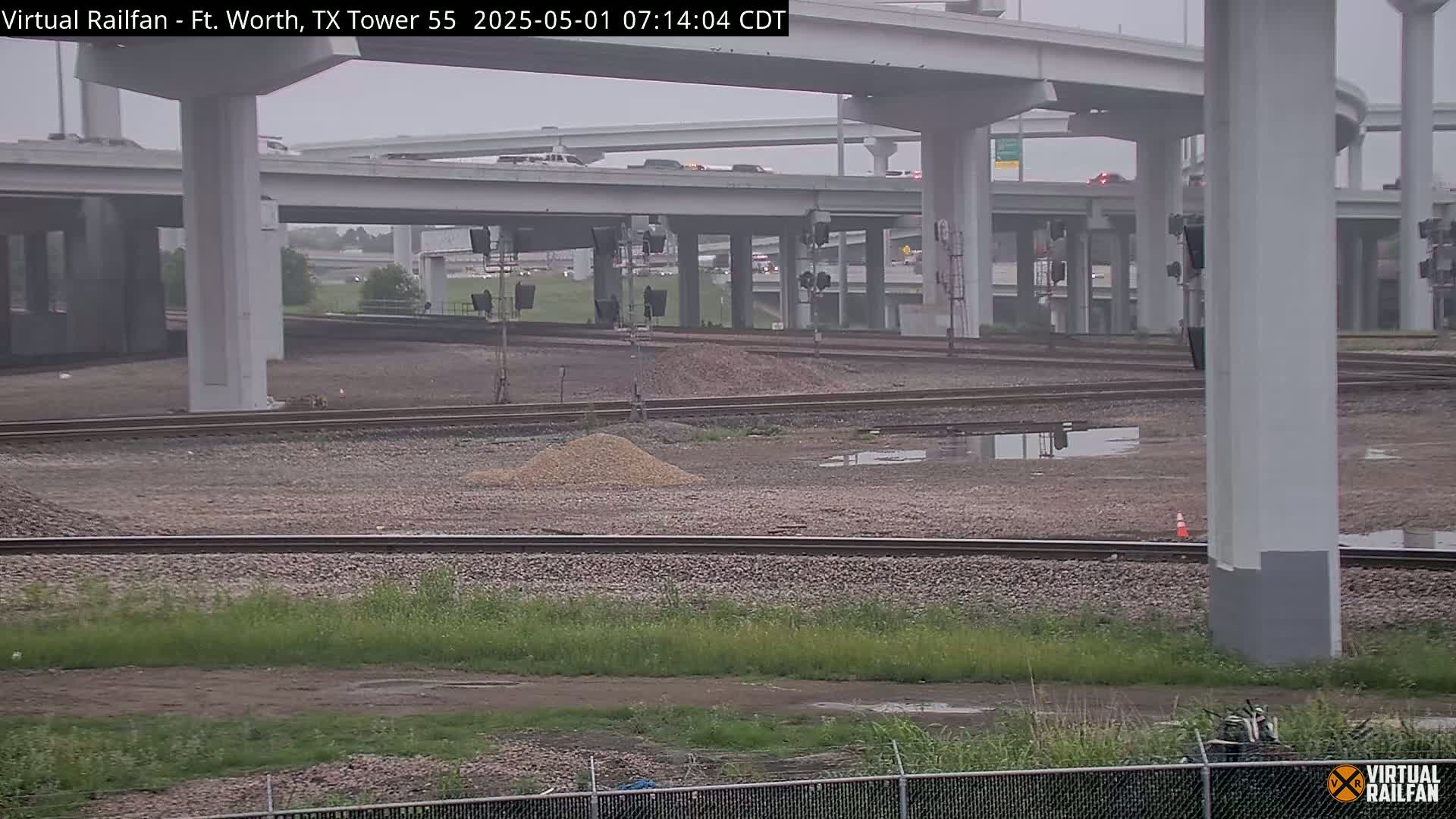 A view of a railway switching yard next to a highway overpass on an overcast day, with puddles in the ground.