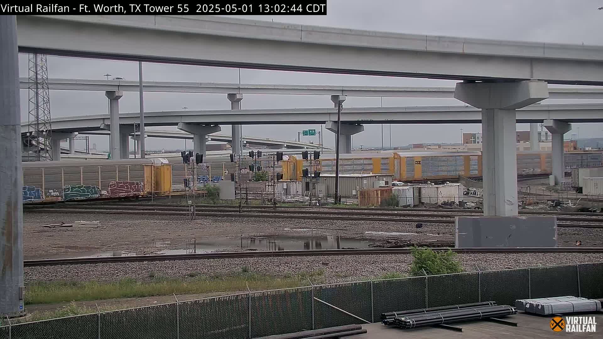 A view of railway tracks and freight cars under an overpass on an overcast day, with puddles of water on the ground.