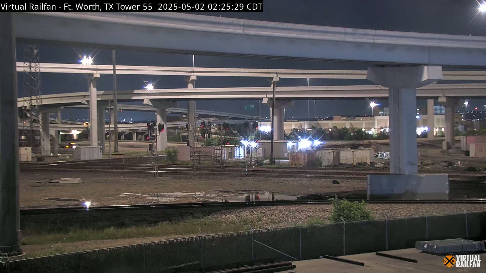 At night, under a dark sky, a nighttime view shows railroad tracks and a large highway overpass with many pillars and levels.