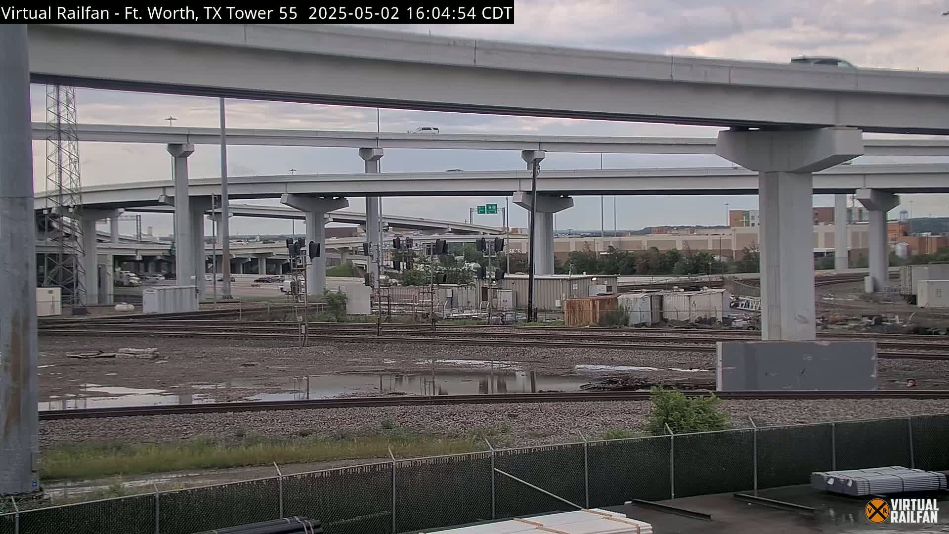 A view of a railroad track with muddy puddles, under a network of elevated highways on an overcast day.