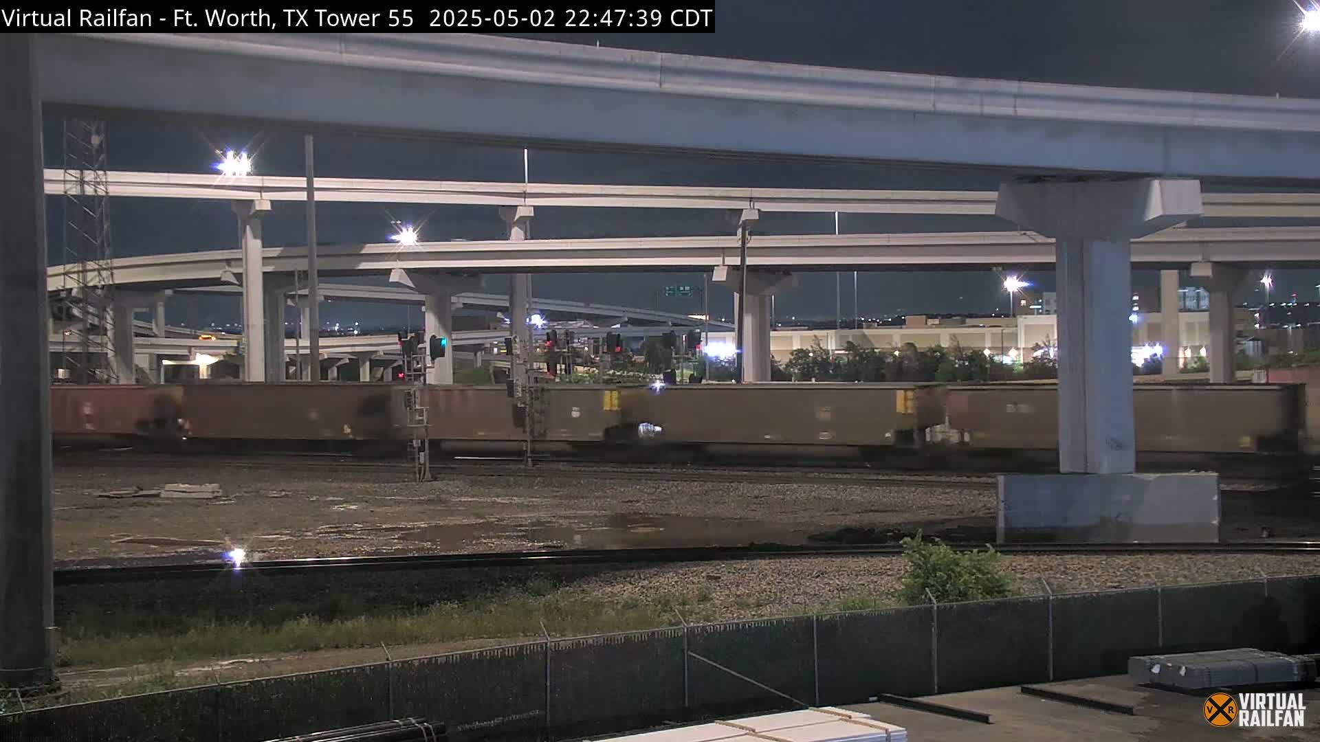 A nighttime view of a freight train moving under a large highway overpass.