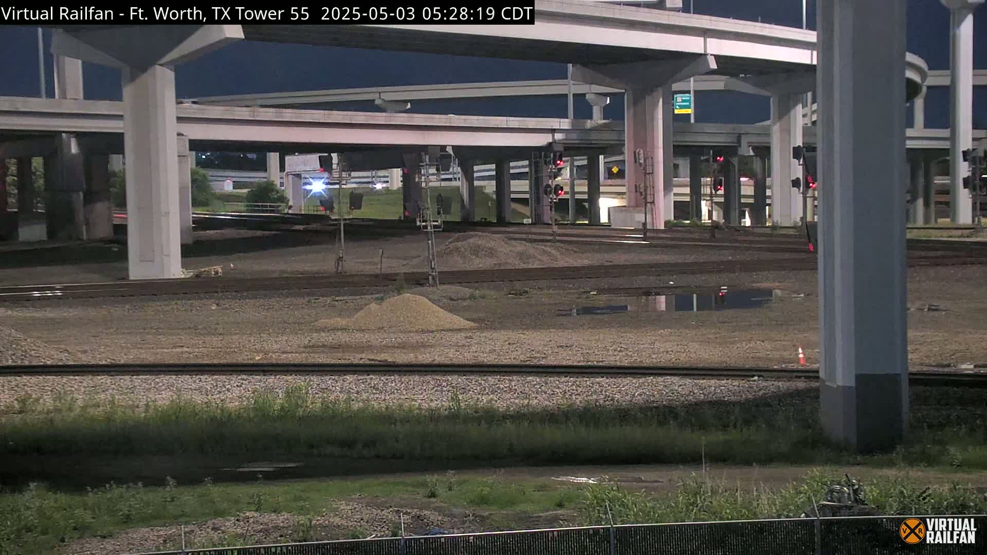 Under a dark night sky, train tracks run beneath a large highway overpass, with some construction materials and puddles visible on the ground.