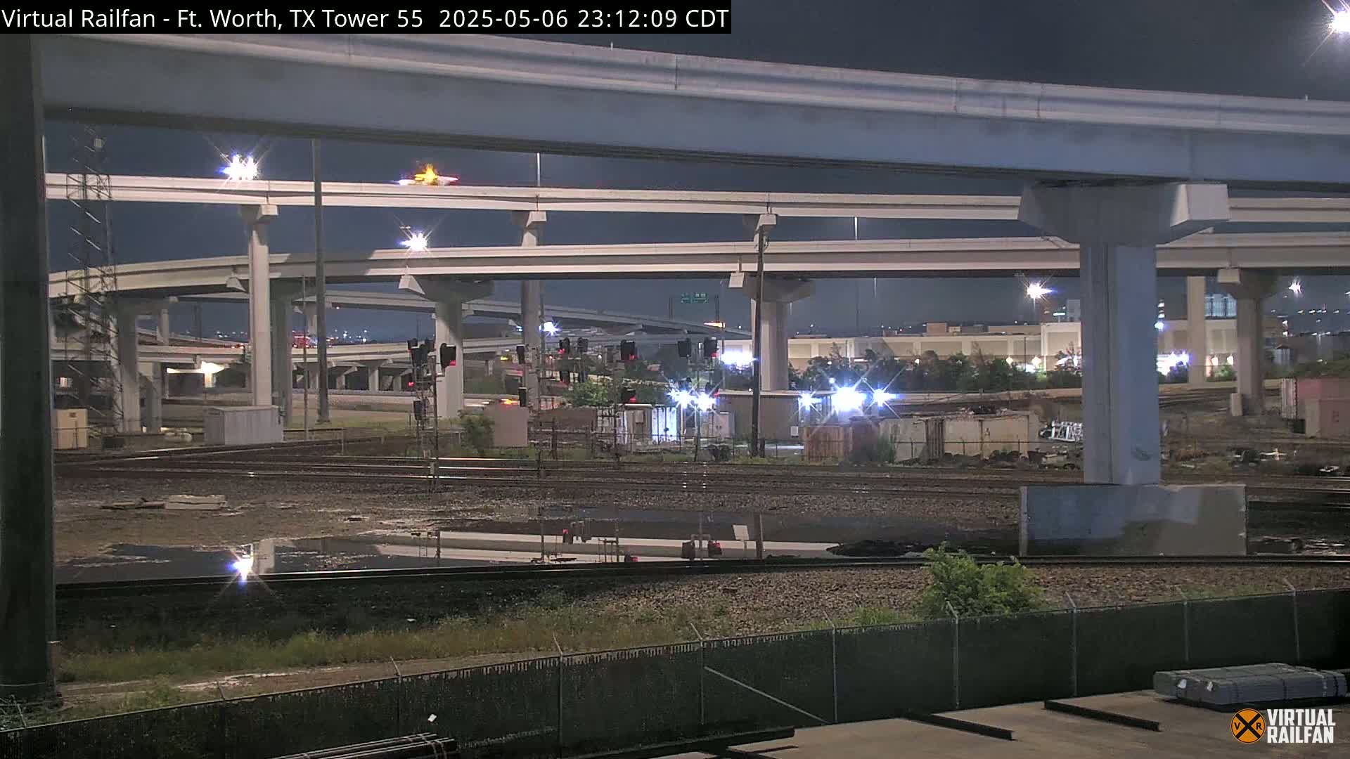 At night, under a clear sky, a view shows a complex highway overpass system above railroad tracks and some industrial buildings.