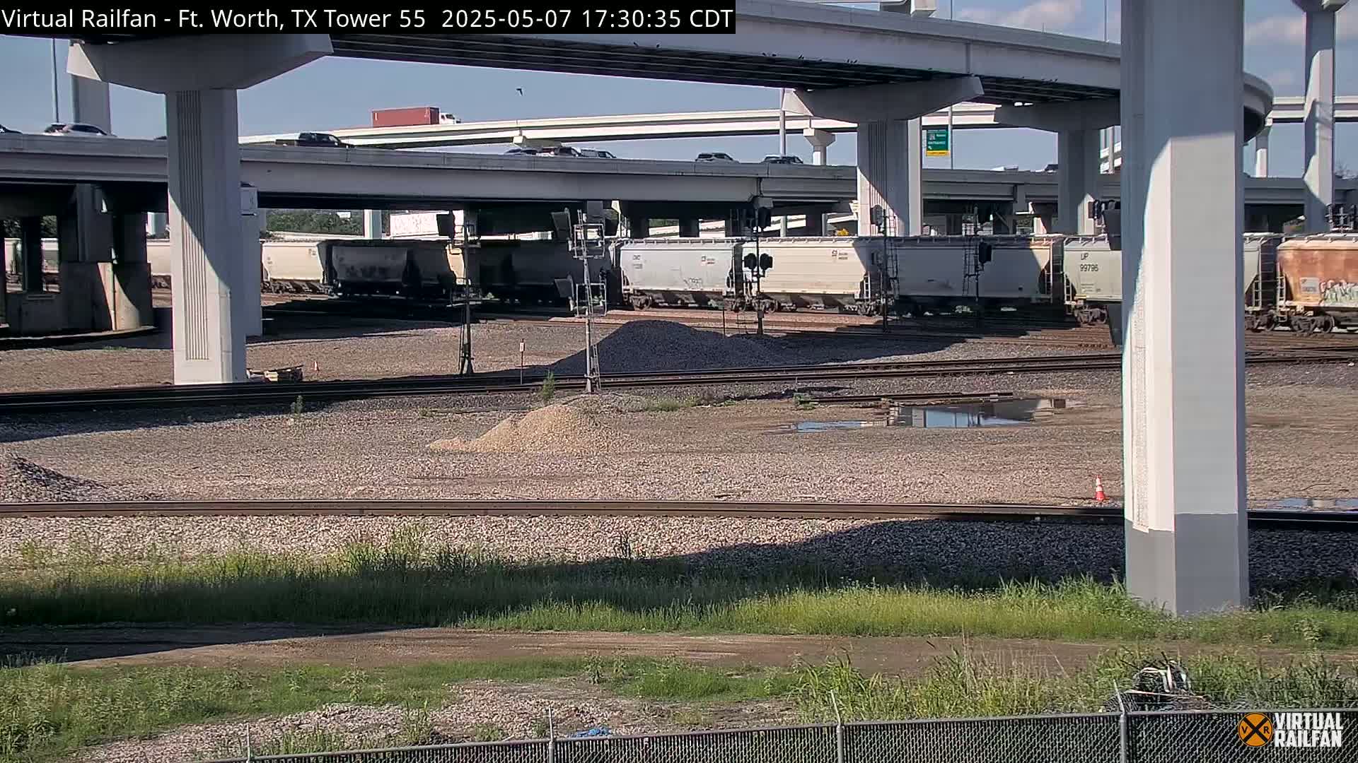 A train of freight cars sits on railway tracks below a highway overpass on a partly sunny day.