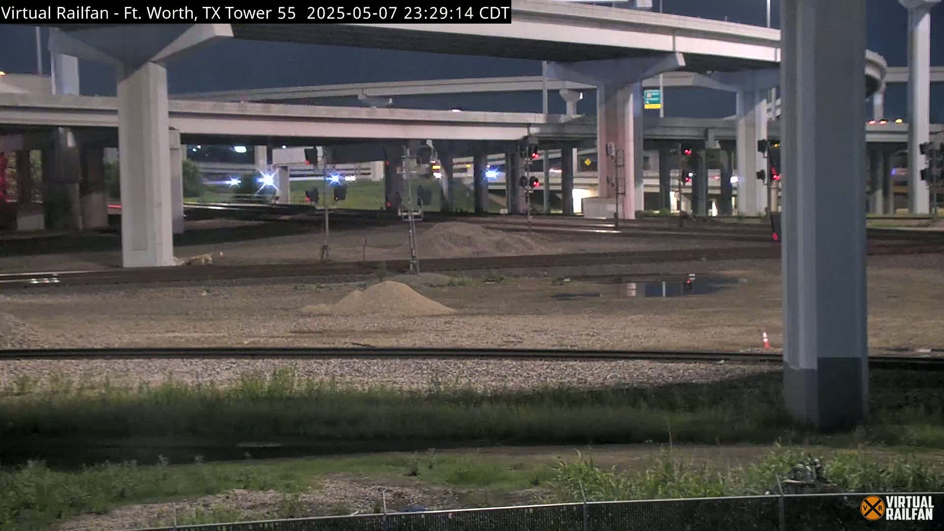 A nighttime view of train tracks beneath an elevated highway interchange, with some small piles of dirt and sparse vegetation.