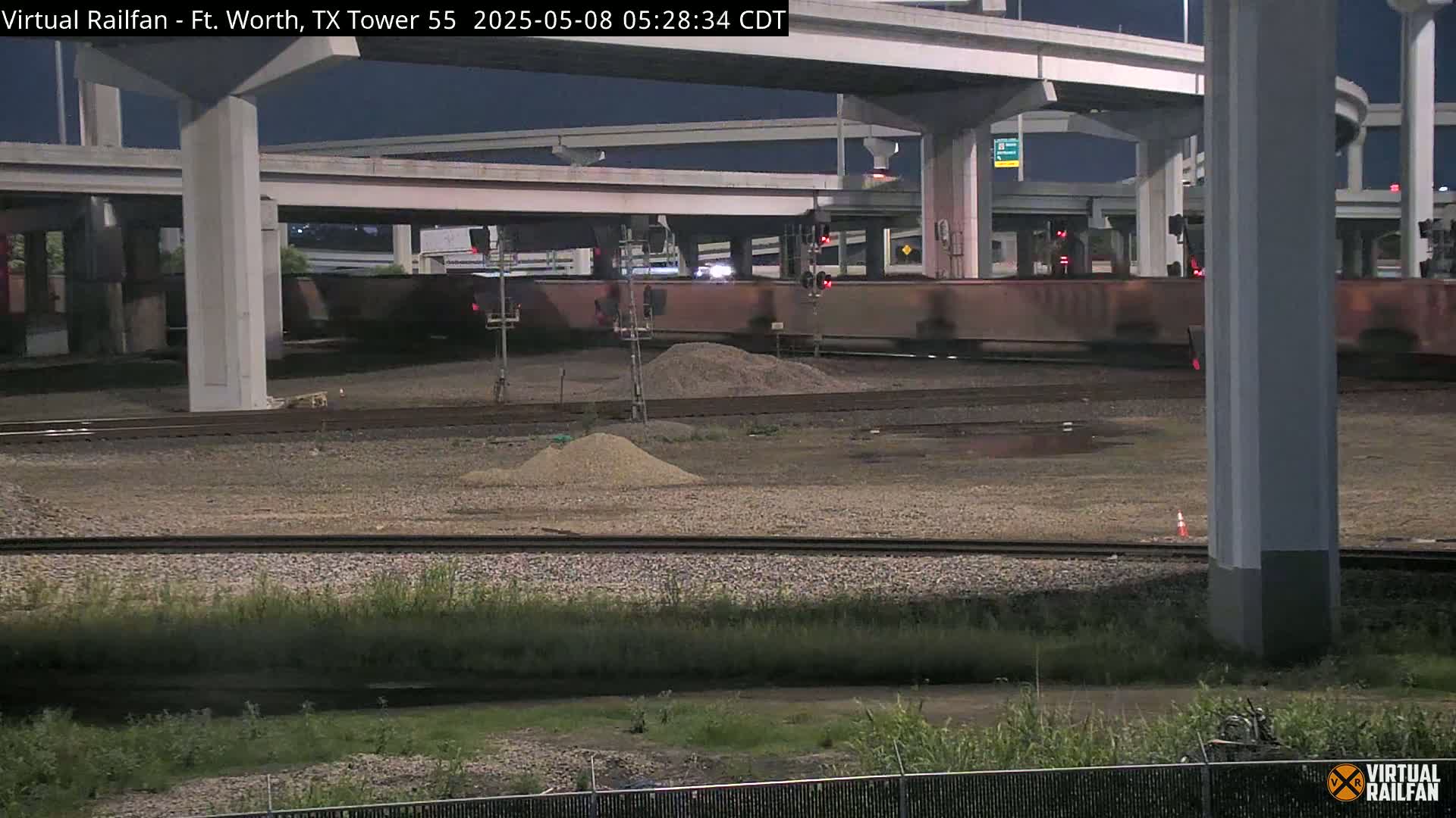 At night, a freight train passes under a highway overpass near a construction site with piles of gravel.