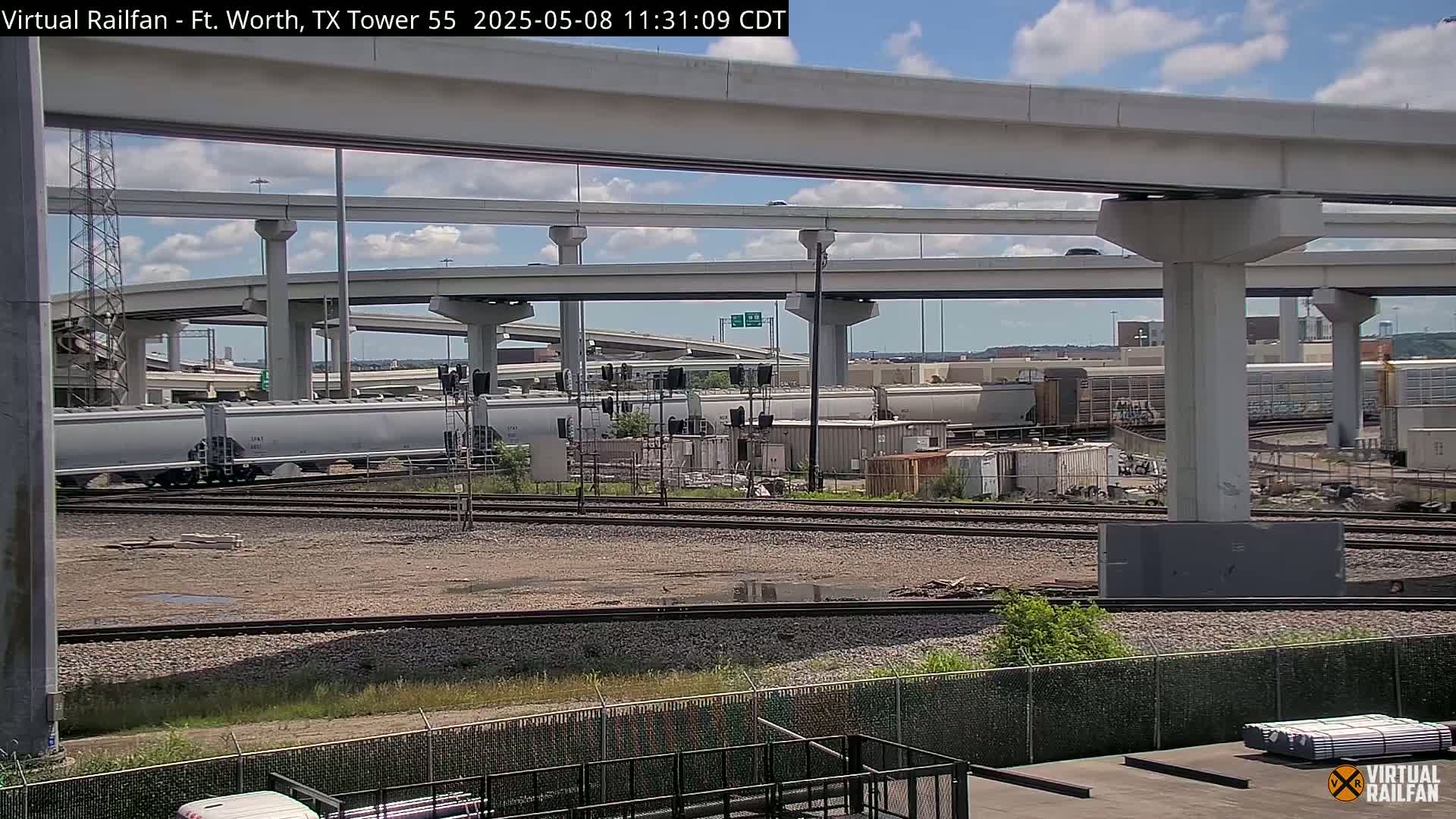 Railroad tracks and a large elevated highway overpass are visible under a partly cloudy sky.