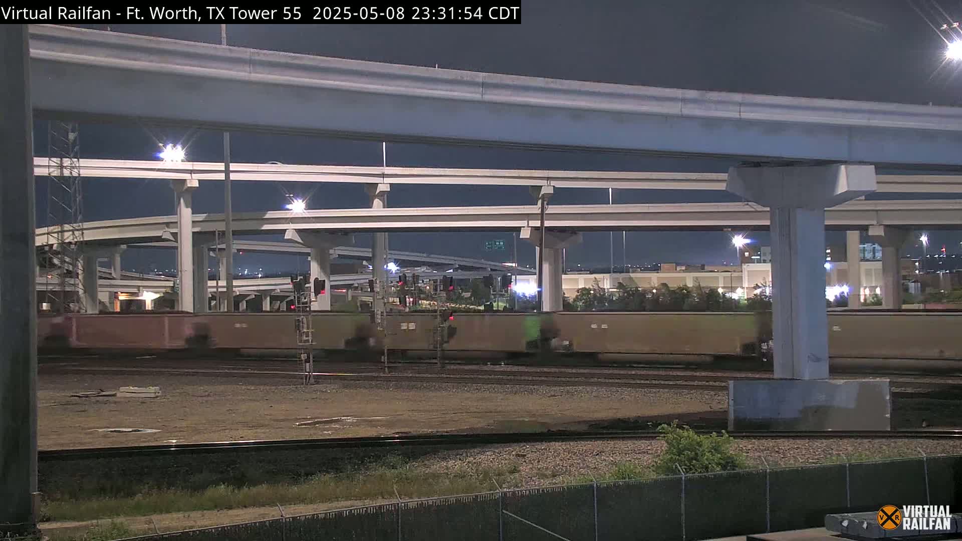 A freight train passes under a large highway overpass at night.