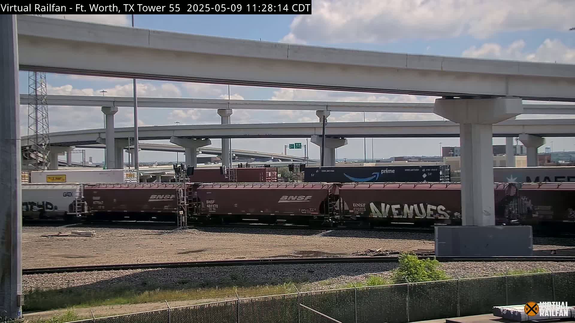 A section of railroad tracks with several maroon freight cars sits below a large highway overpass on a partly cloudy day.