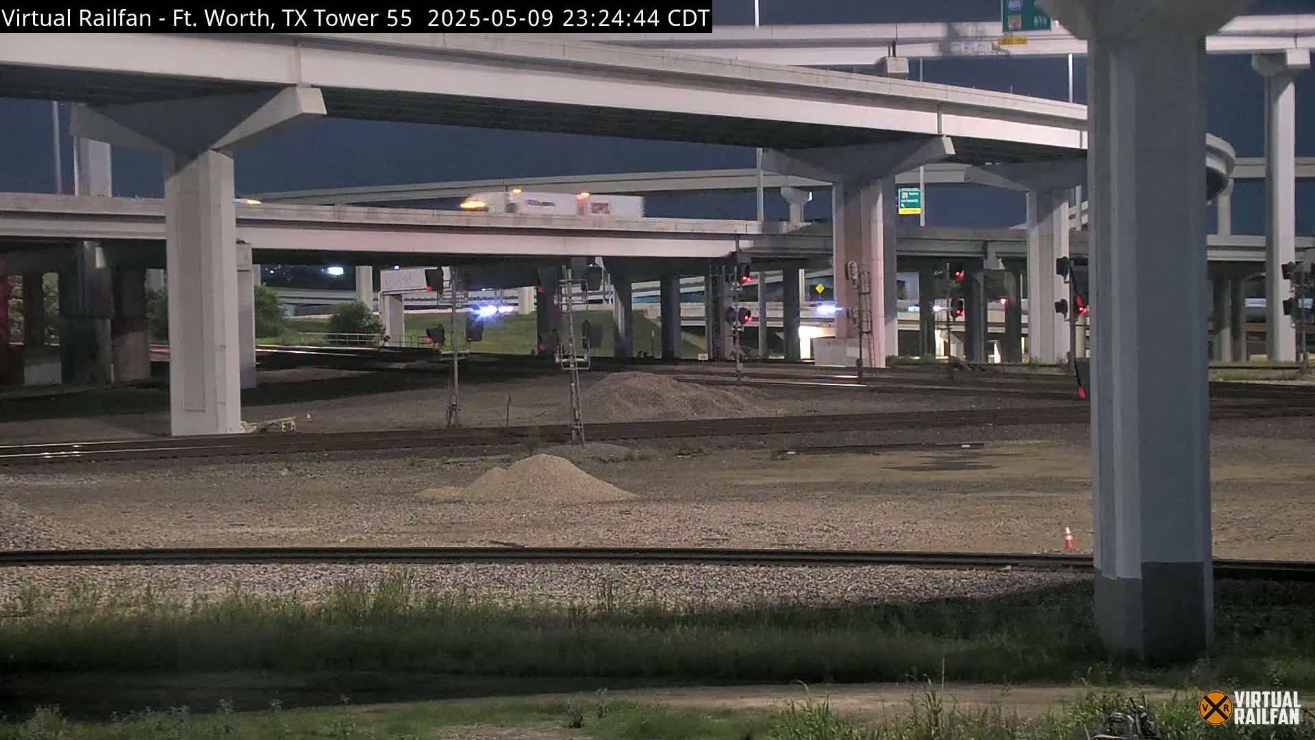Under a dark night sky, railroad tracks and a highway overpass intersect, with piles of gravel and minimal vegetation visible.
