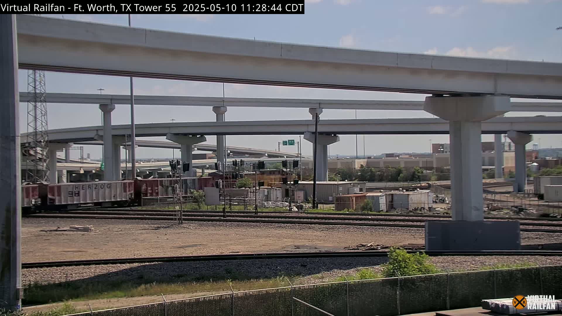 A freight train passes under a complex network of elevated highway overpasses on a partly sunny day.