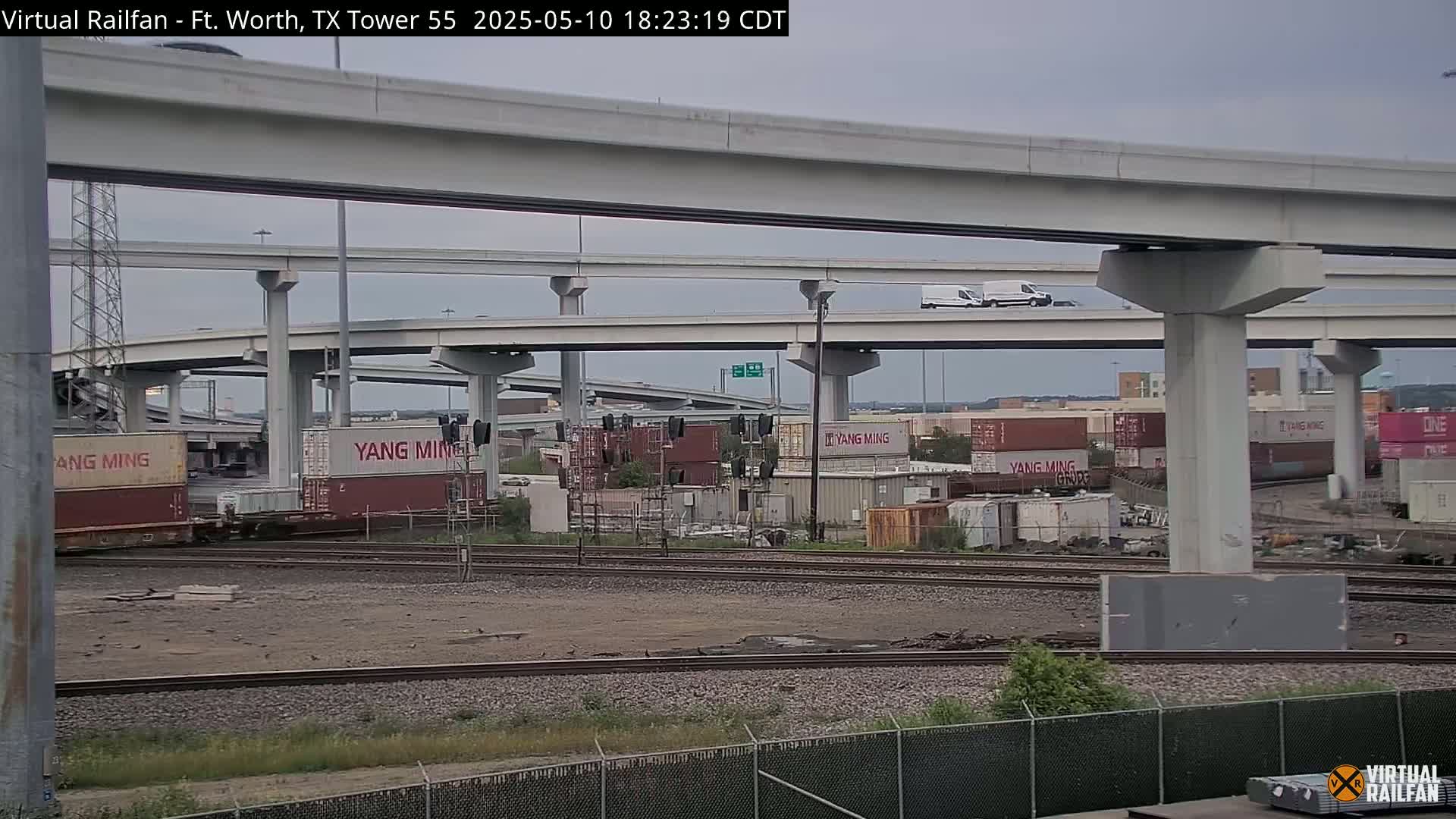 A view of a railroad yard with shipping containers and a complex highway overpass system under an overcast sky.