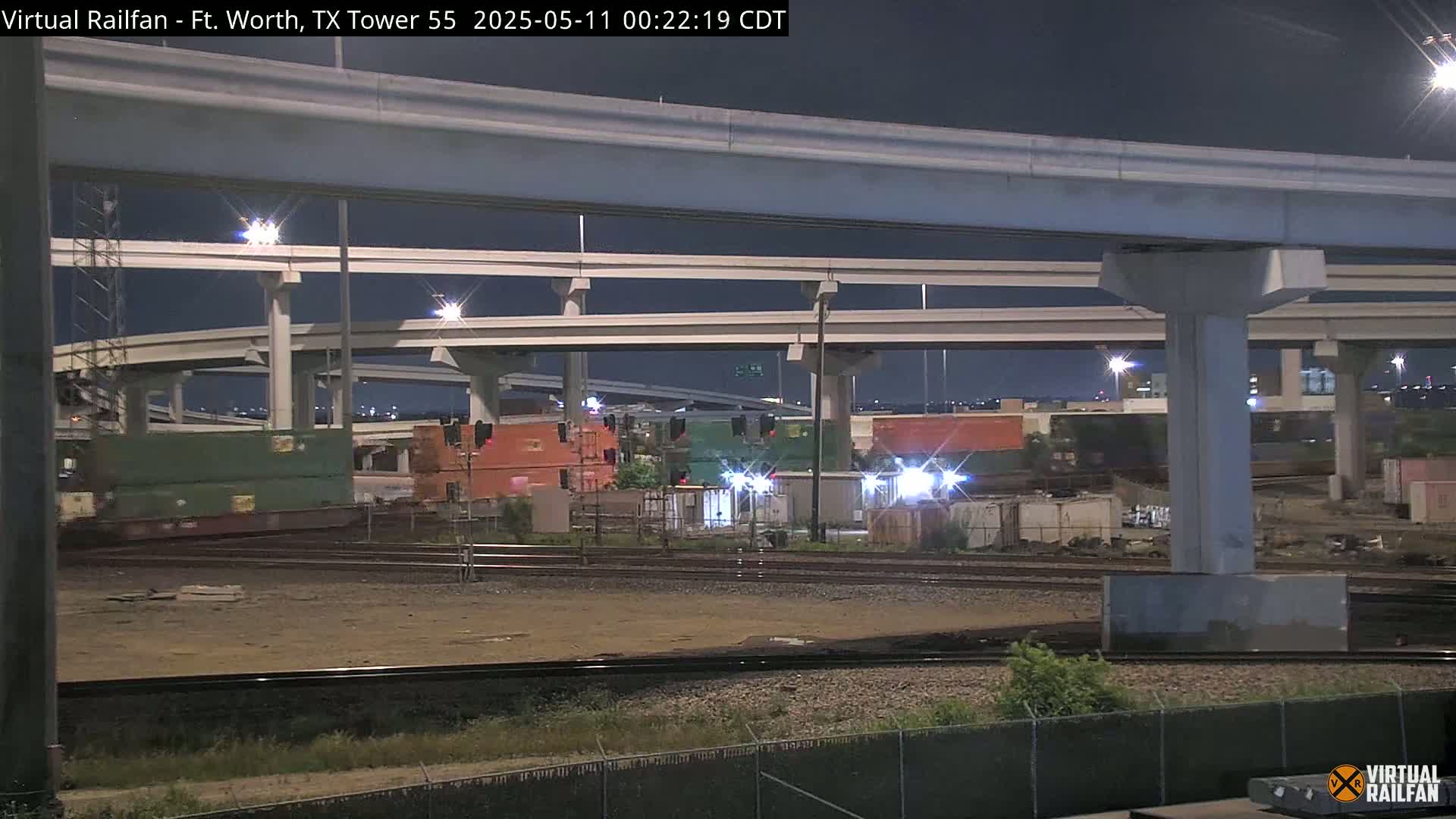 At night, a freight train sits under a large highway overpass.
