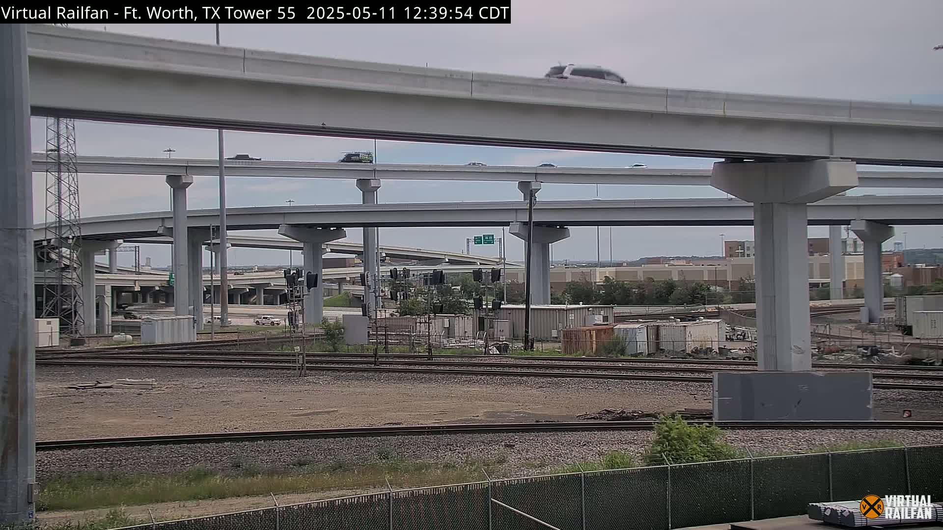 Under an overcast sky, several layers of highway overpasses and supporting pillars are seen over train tracks and a fenced-off area with various containers and debris.