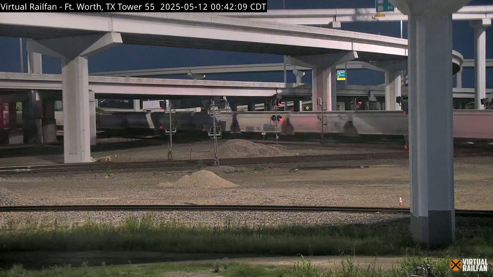 A nighttime view of a freight train passing under a highway overpass.